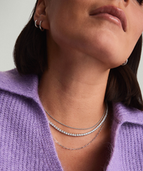 Close-up of a person wearing a Lab Grown Sapphire Tennis Necklace, featuring clear stones set in silver, layered with two other silver chains and silver hoop earrings.