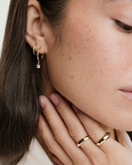 Close-up of a woman's ear wearing Round Lab Grown Sapphire Hoops, featuring one plain gold hoop and one with a dangling stone, complemented by gold rings on her hand.