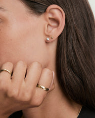 Side-profile of a woman's ear adorned with Round Lab Grown Sapphire Studs alongside a small gold ball, with gold rings on her hand.