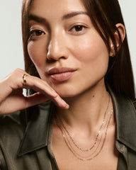 Young woman wearing the Mini Anchor Chain Necklace, featuring interconnected links, layered with delicate chains, complemented by simple gold jewelry. Hand rests on chin.