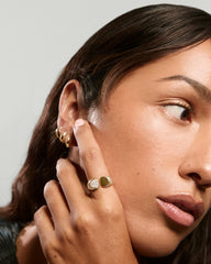 Woman wearing the Sabine Open Signet Pinky Ring, featuring a smooth gold band and pavé diamond cluster, gently touching her ear adorned with gold hoops.