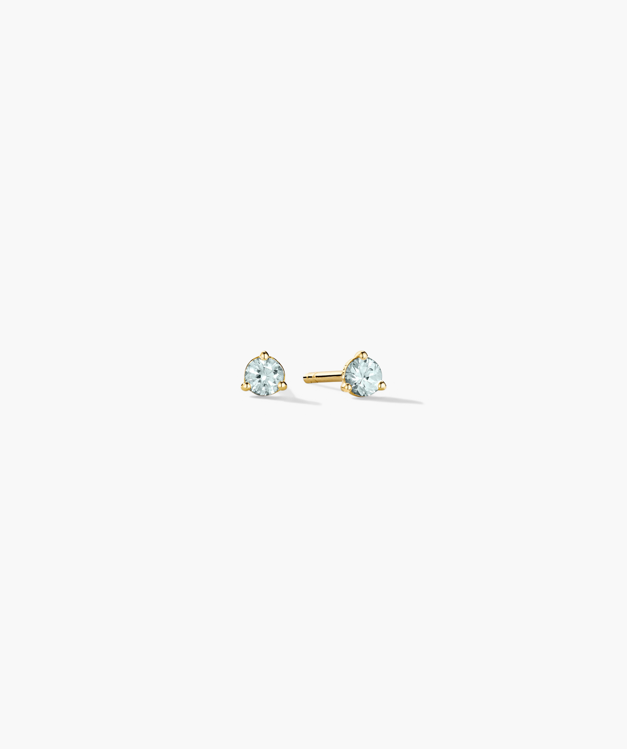 Gemstone Mini Studs featuring round, light-blue faceted stones in three-prong yellow-gold settings; one earring faces forward, the other shows the post.