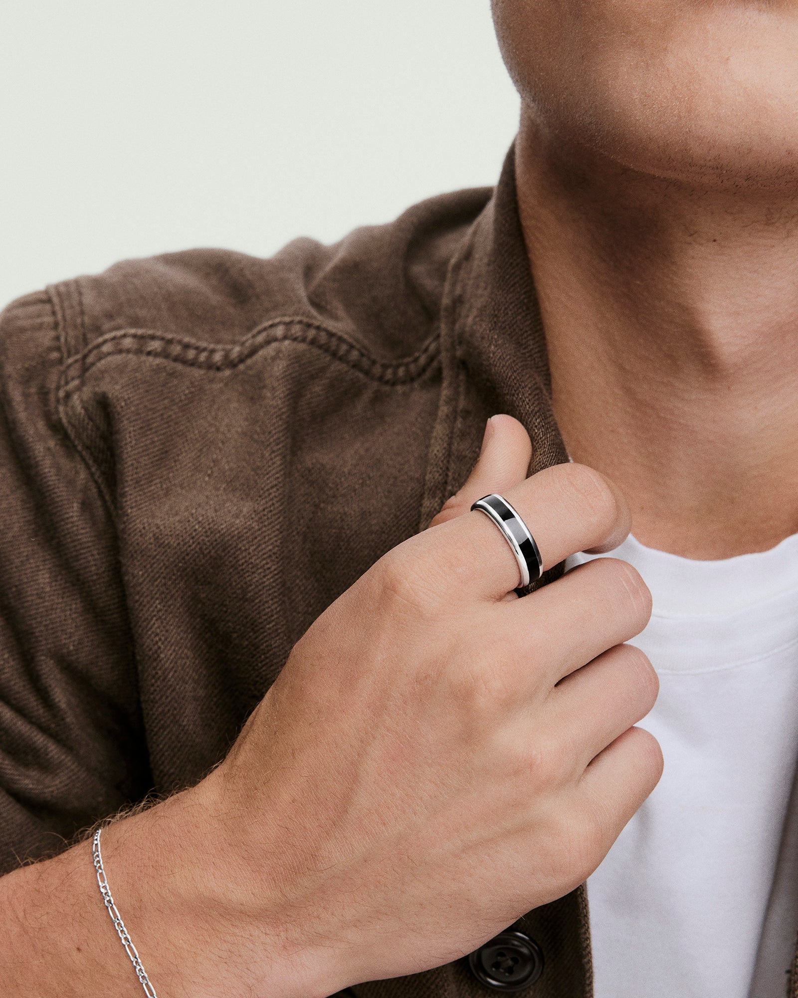 A hand wearing the Band Ring Black Enamel Inlay on the middle finger, complemented by a thin silver chain bracelet, against a white T-shirt and brown jacket.