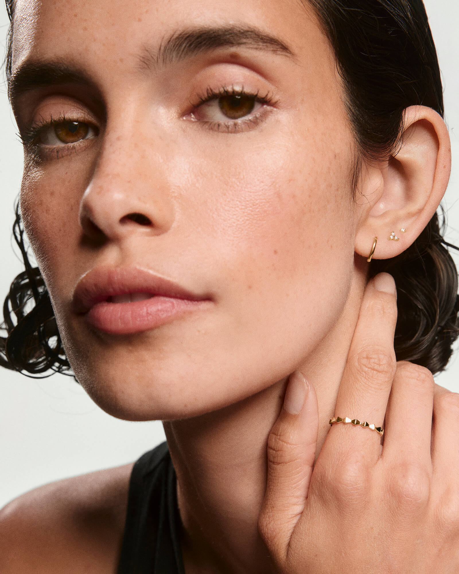 Woman wearing the Sculpture Ring, a slim gold band, on her right hand, complemented by gold earrings, against her lightly freckled face.