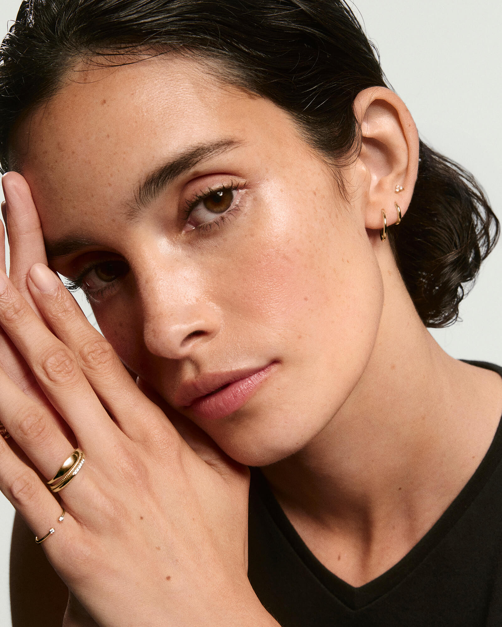 Young woman with slicked-back hair wearing Mini Lotus Diamond Studs, alongside slim gold rings and small hoop earrings, against a neutral background.