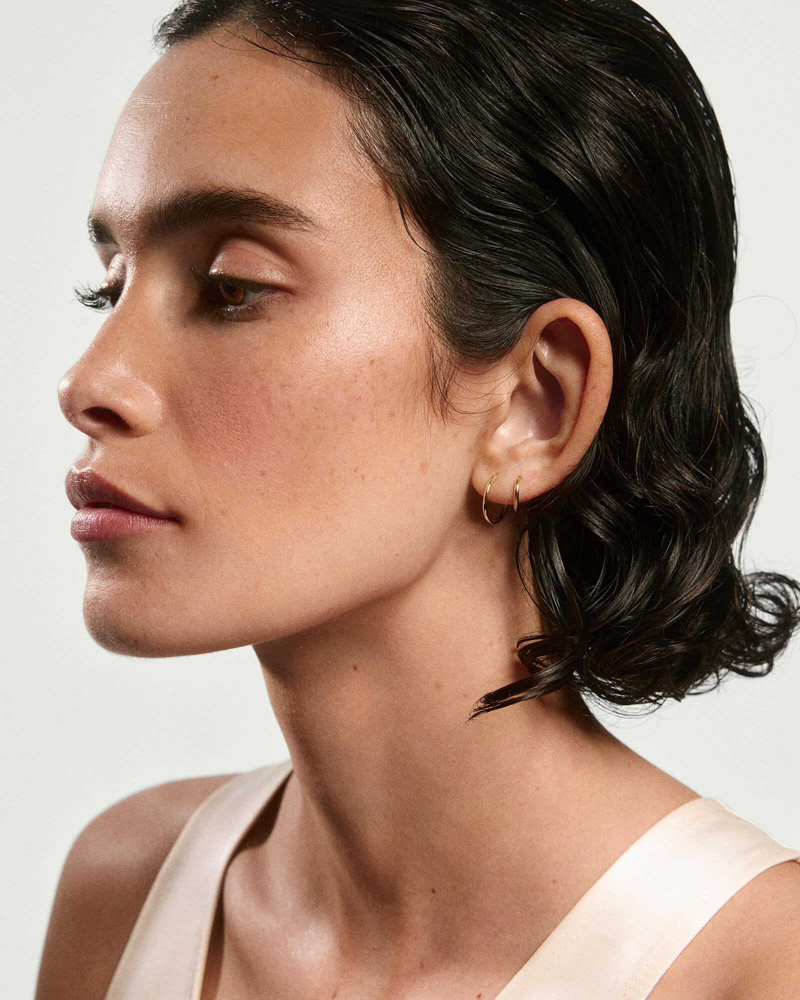 Side-profile of a person wearing Small Hoops, the lightest gold earrings, with slicked-back hair, minimal makeup, and a sleeveless top against a plain background.