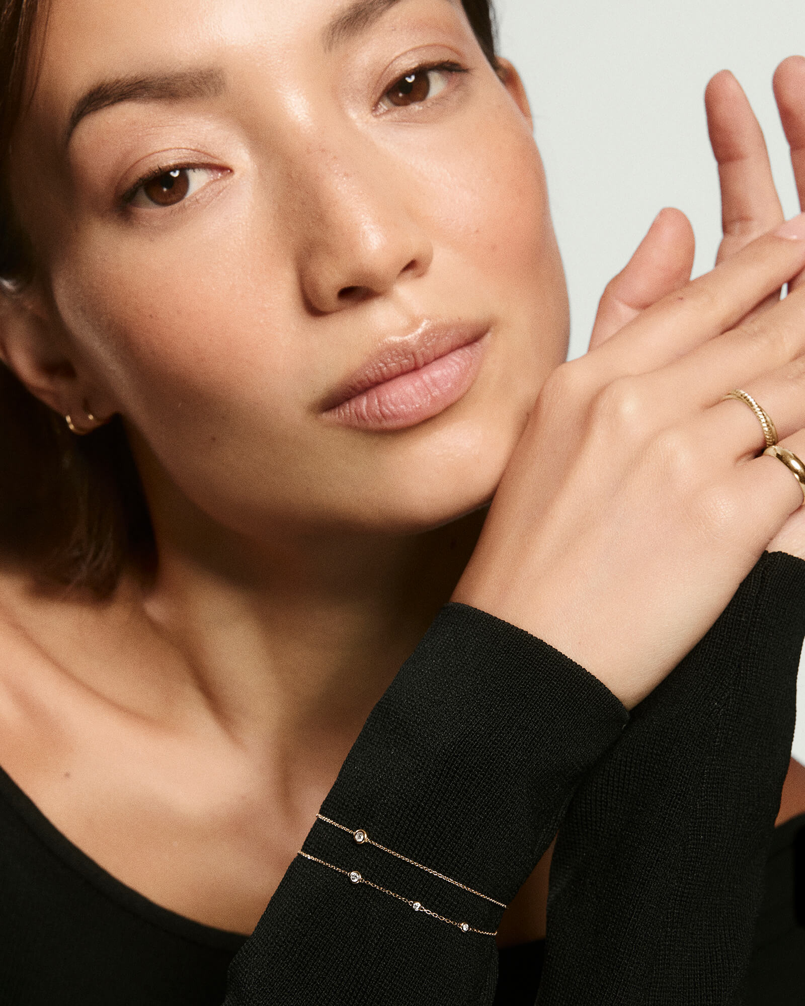 Woman's hands framing her face, showcasing the Solo Diamond Braceletâa fine gold chain with a bezel-set, round-cut diamond, complemented by simple gold rings.