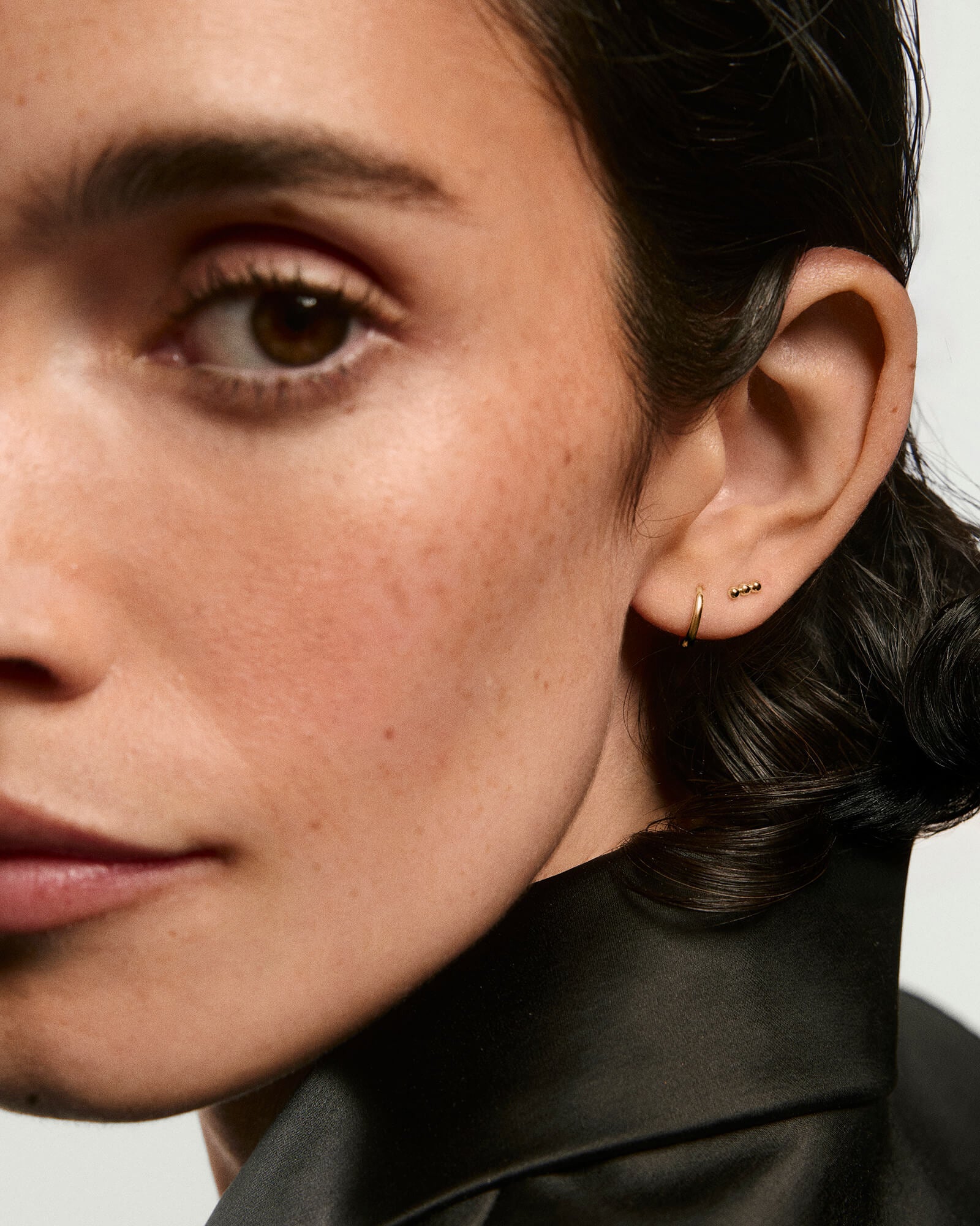 Close-up of a woman wearing Spheres Bar Studsâa trio of small gold ball studsâpaired with a tiny gold hoop, highlighting the earrings' elegance.