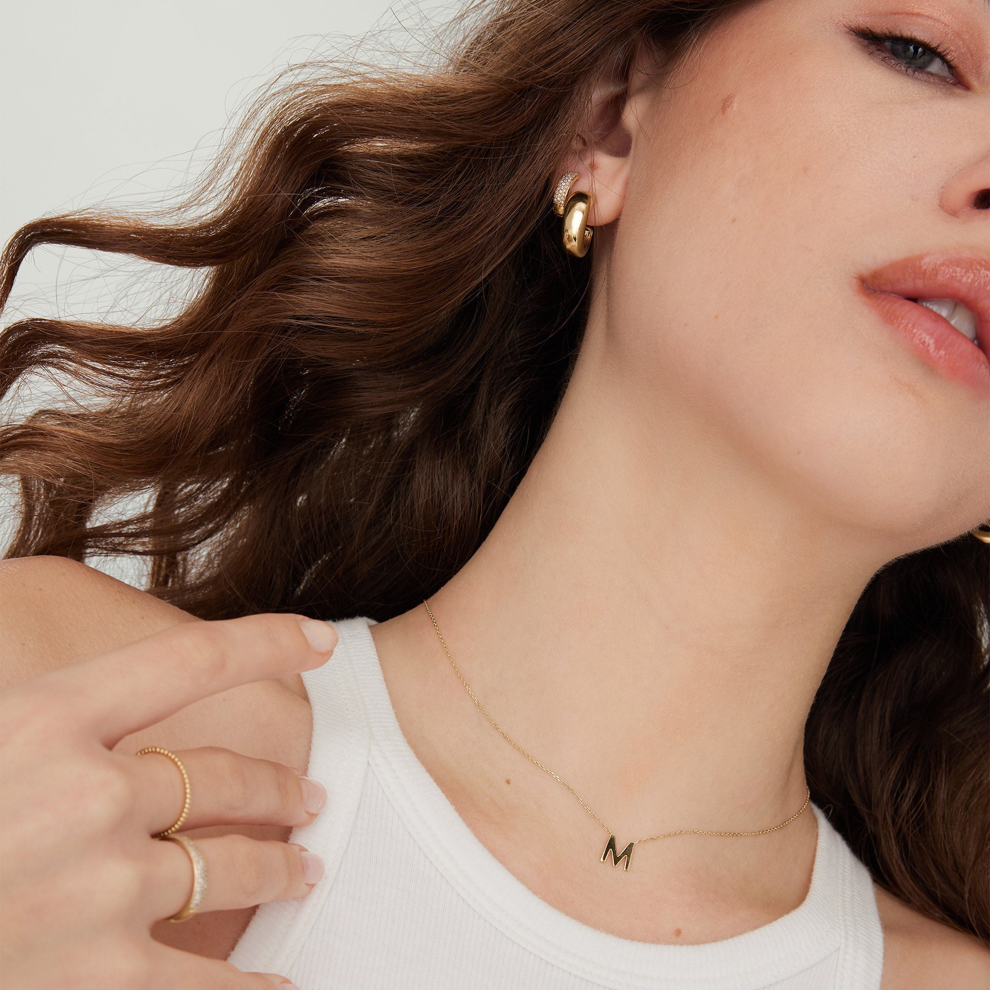 Woman with long brown hair wearing a white tank top, showcasing a gold initial letter necklace.