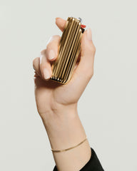 Hand with gold bracelet holds Charlotte Lighter Case, a ribbed-metal gold lighter. Thumb is near the ignition wheel, showcasing its elegant design.