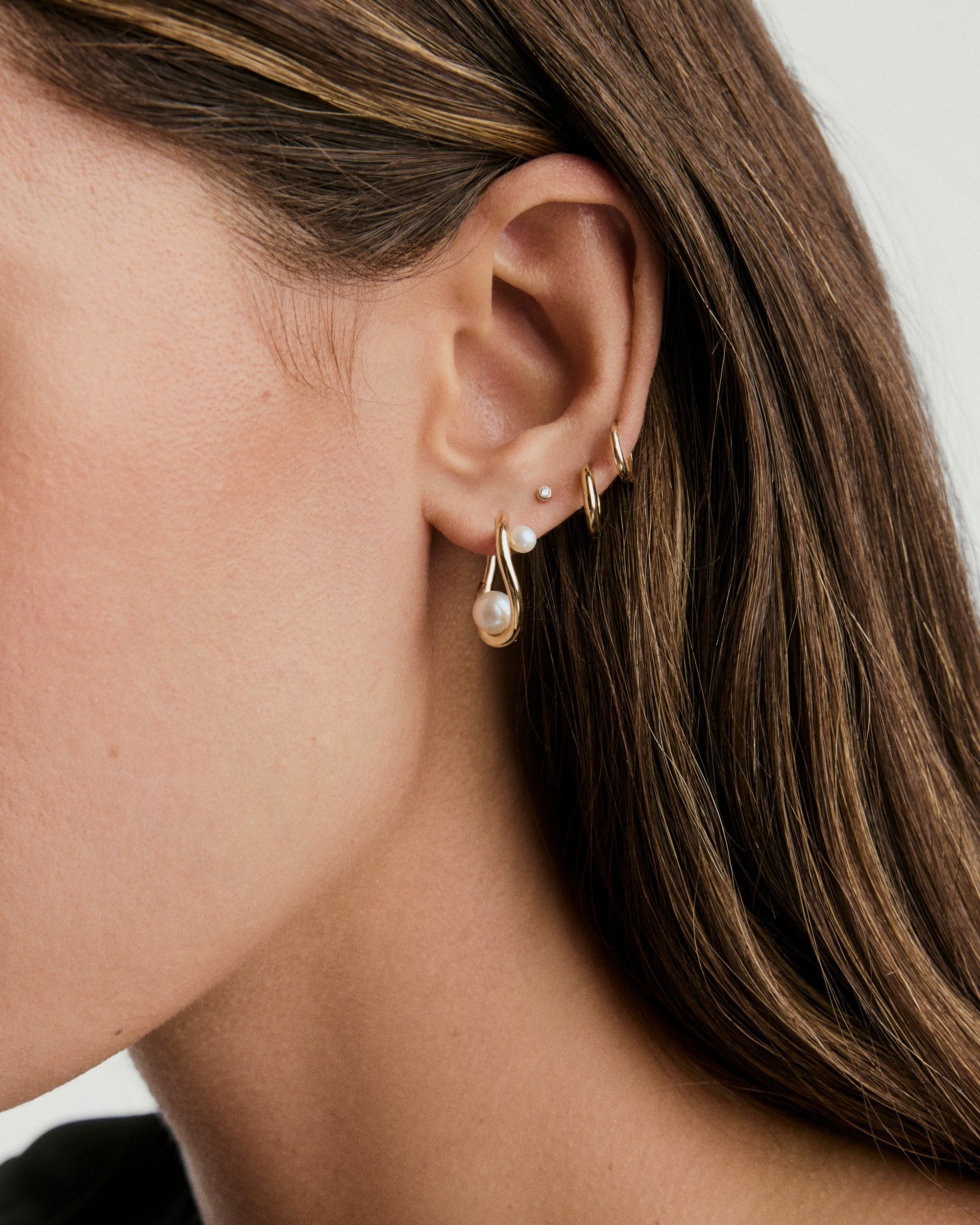 Close-up of a woman's ear wearing DÃ´me Figure Pearl Hoops, showcasing interwoven pearls of varying sizes in gold hoop earrings.