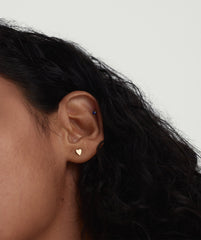 Close-up of a woman's ear showcasing a Mini Heart Stud earring, a delicate heart-shaped fashion accessory. Perfect for adding charm to your jewelry collection.