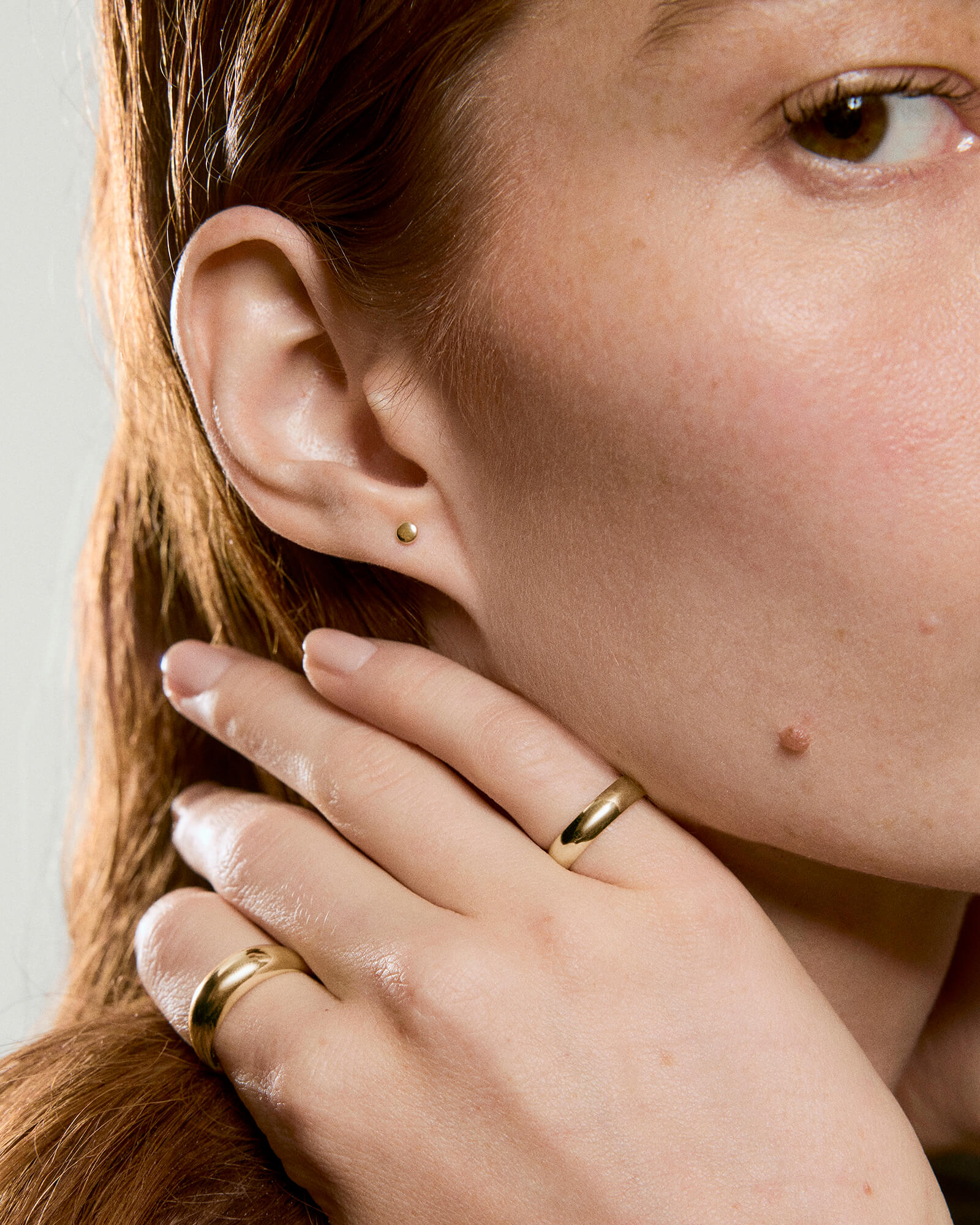 Close-up of a person wearing a Mini Thumbtack Flat Back Stud in gold, highlighting its simple elegance alongside gold rings on fingers.