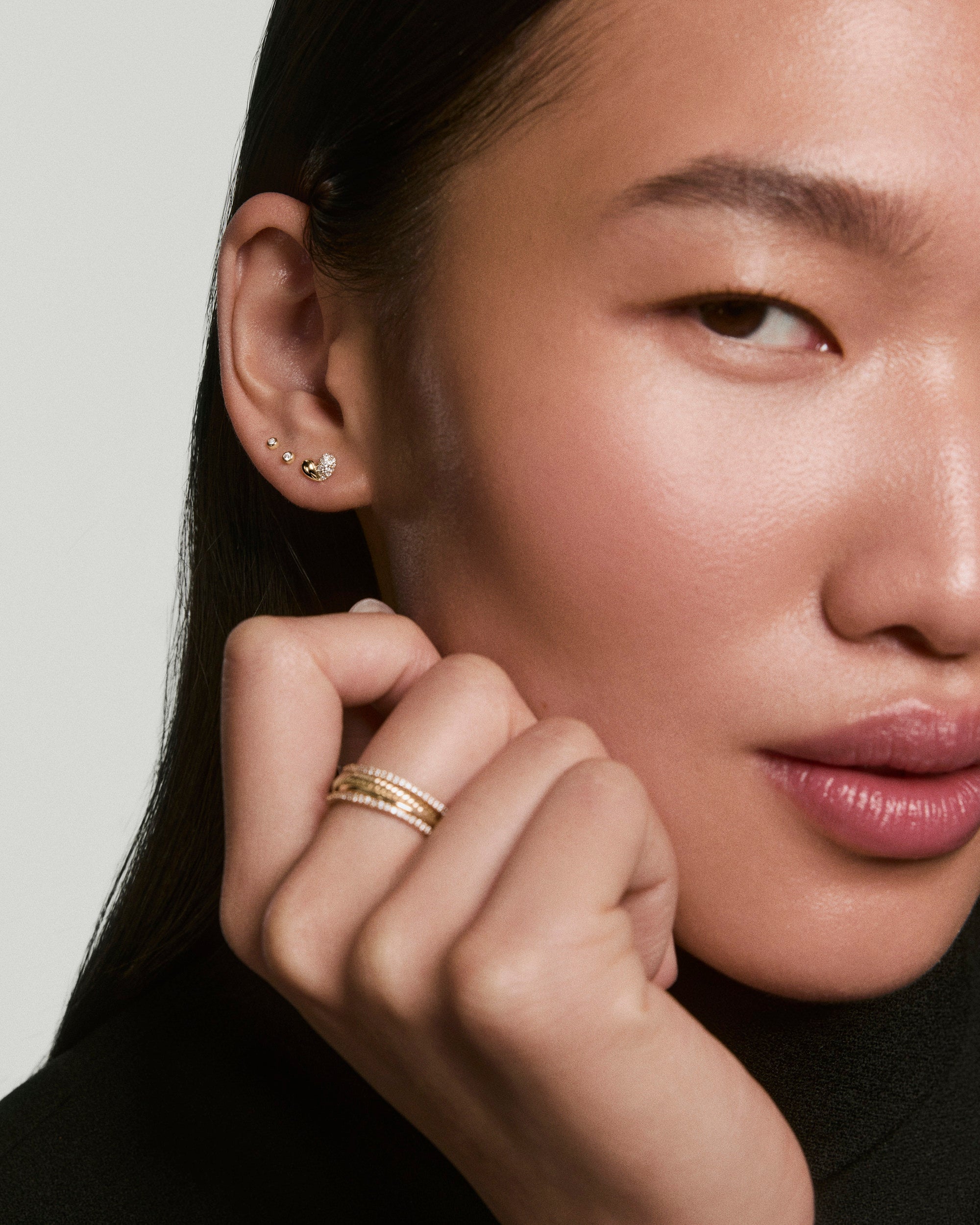 Close-up of a woman's ear wearing Better Half Heart Studs, featuring a tiny heart with a crystal, alongside slim stacked gold rings on her hand.