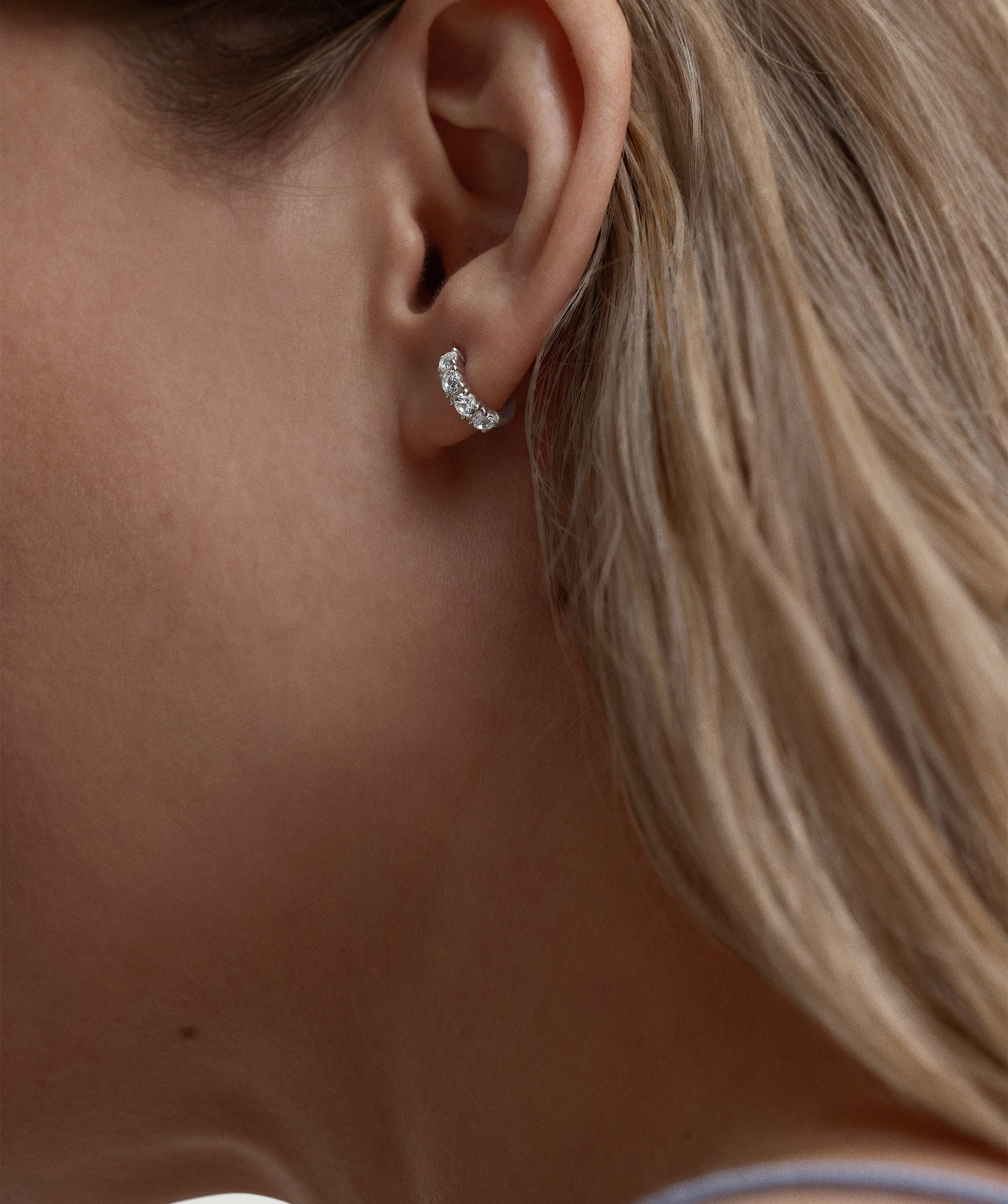 Close-up of a woman's ear wearing Lab Grown Sapphire Huggie earrings, featuring dainty, round white sapphires set in a 12mm hoop.