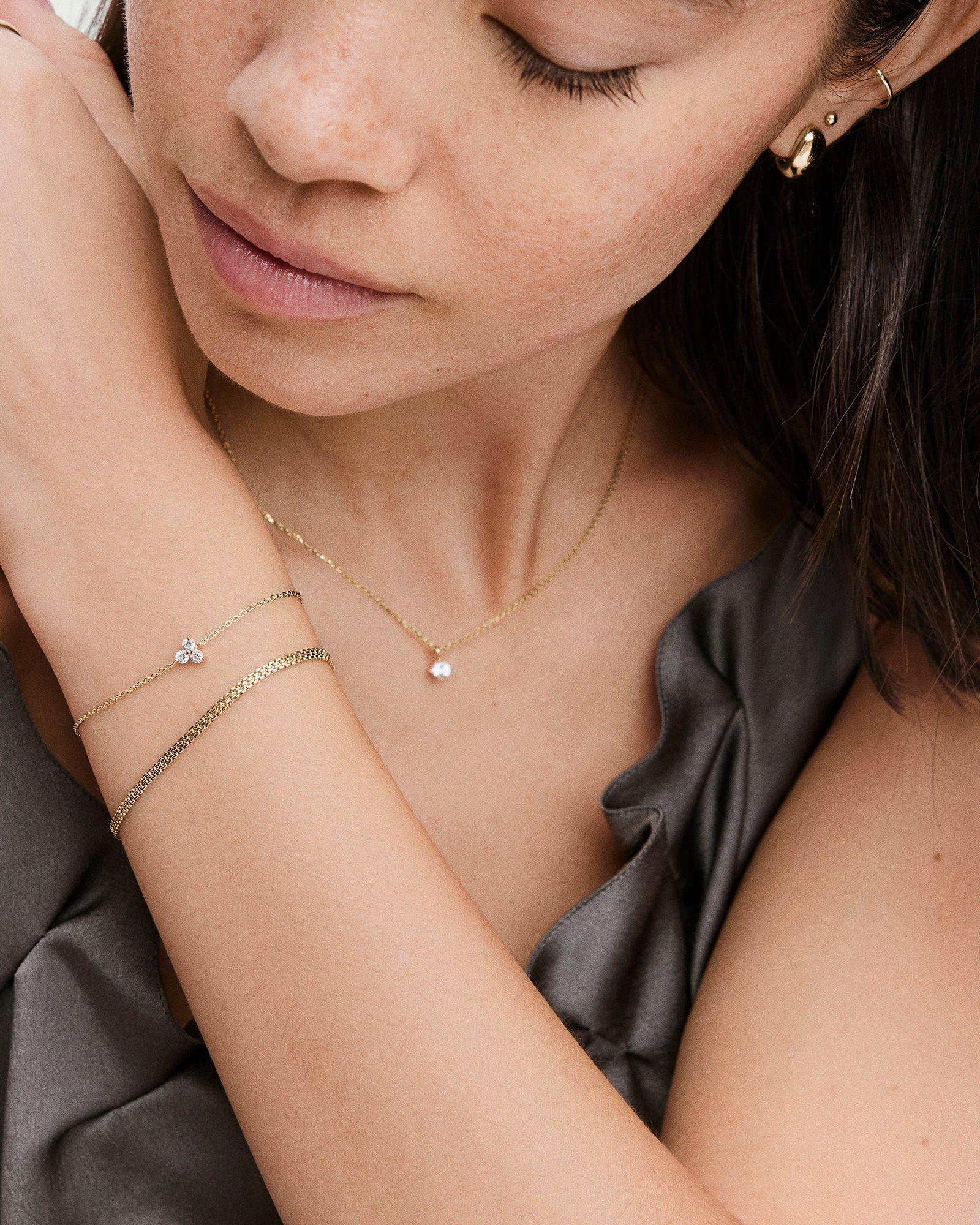 A young woman showcases the Lotus Bracelet, a fine gold chain with three clear stones, alongside a gold necklace and earrings, complementing her satin-like grey top.