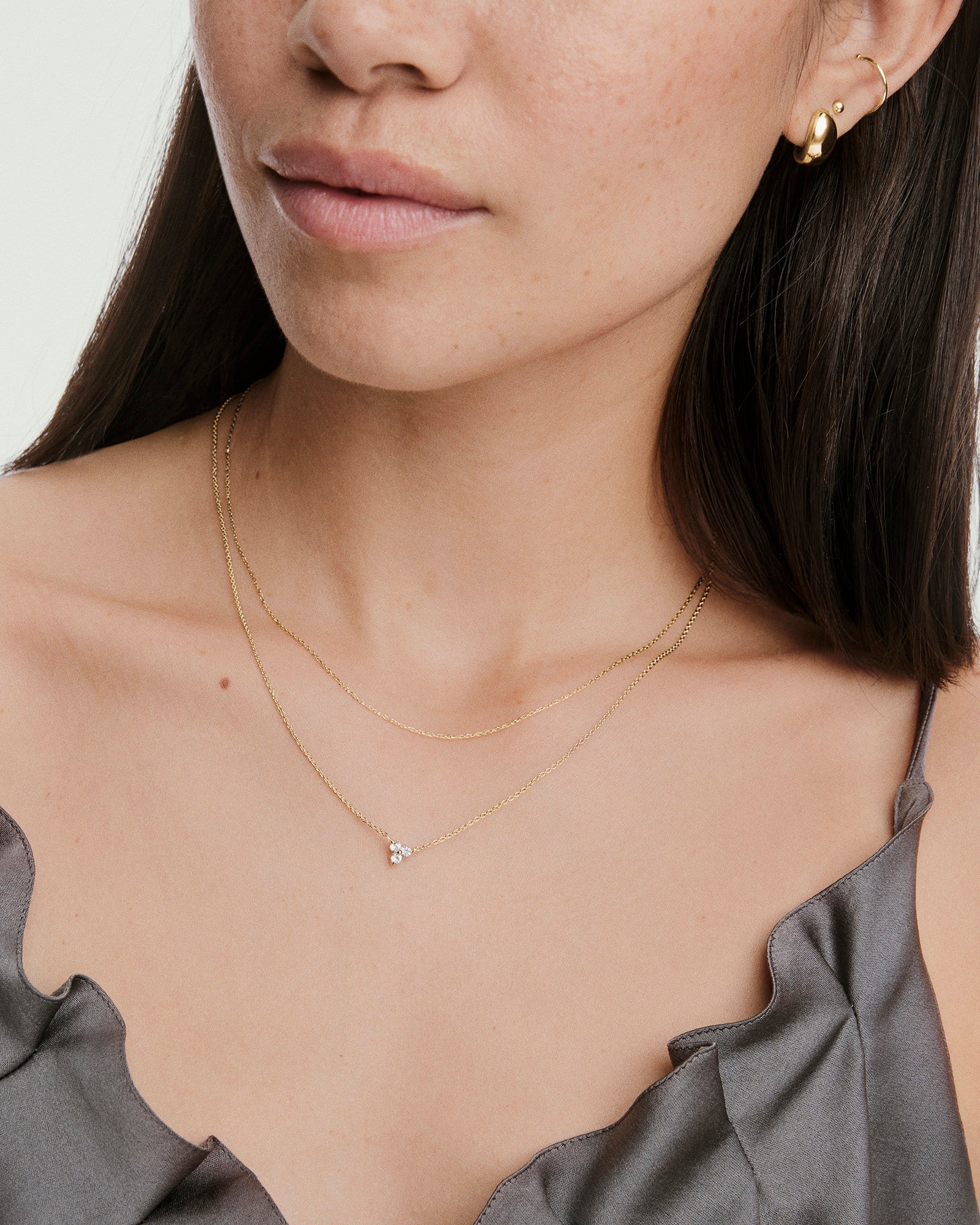 Close-up of a woman's neck wearing the Lotus Necklace, featuring layered gold chains with a small cluster of three white stones, complemented by gold earrings.
