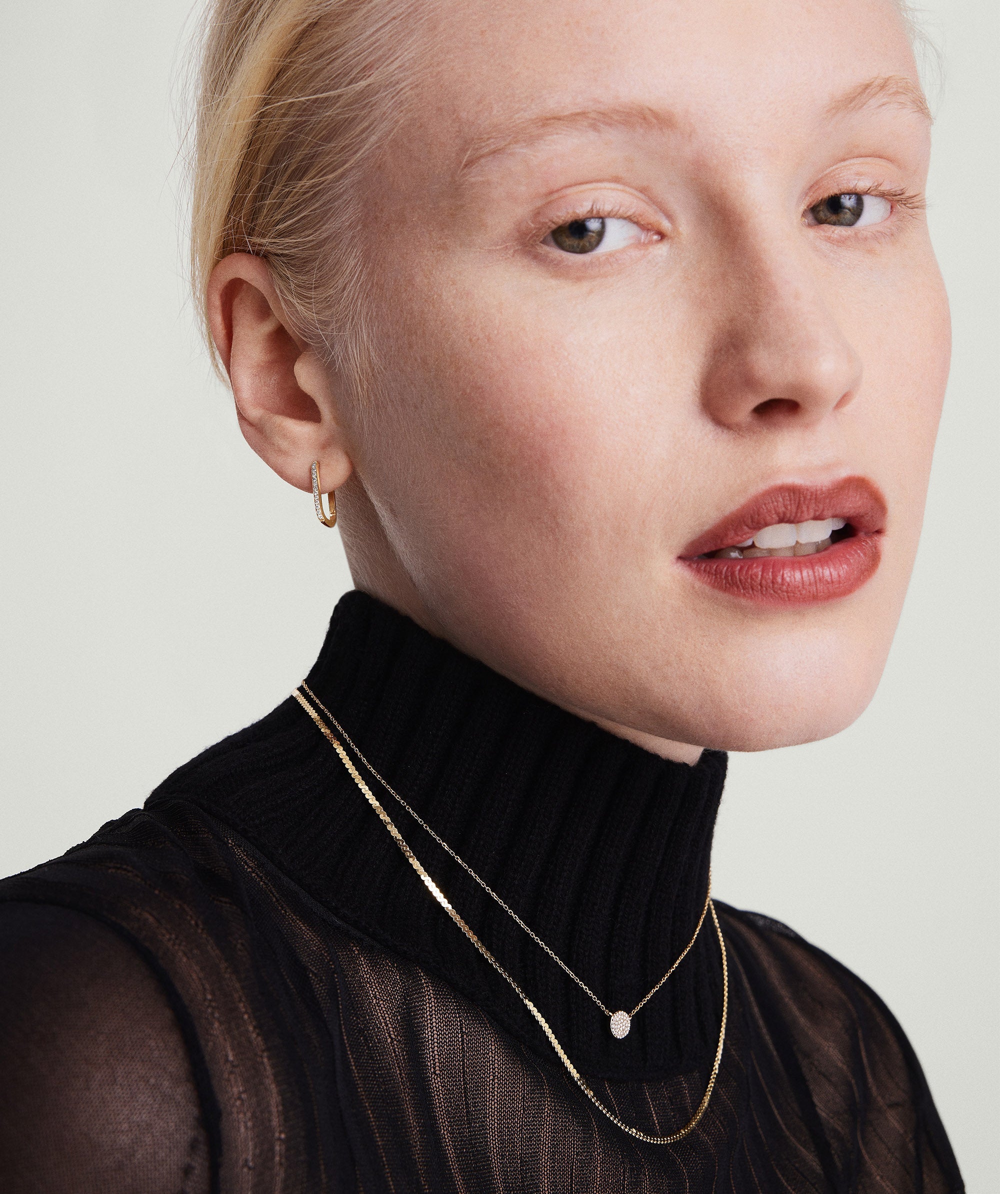 A woman wearing PavÃ© Diamond Small U Hoops, 14k gold earrings set with diamonds, paired with a black turtleneck and gold necklaces.