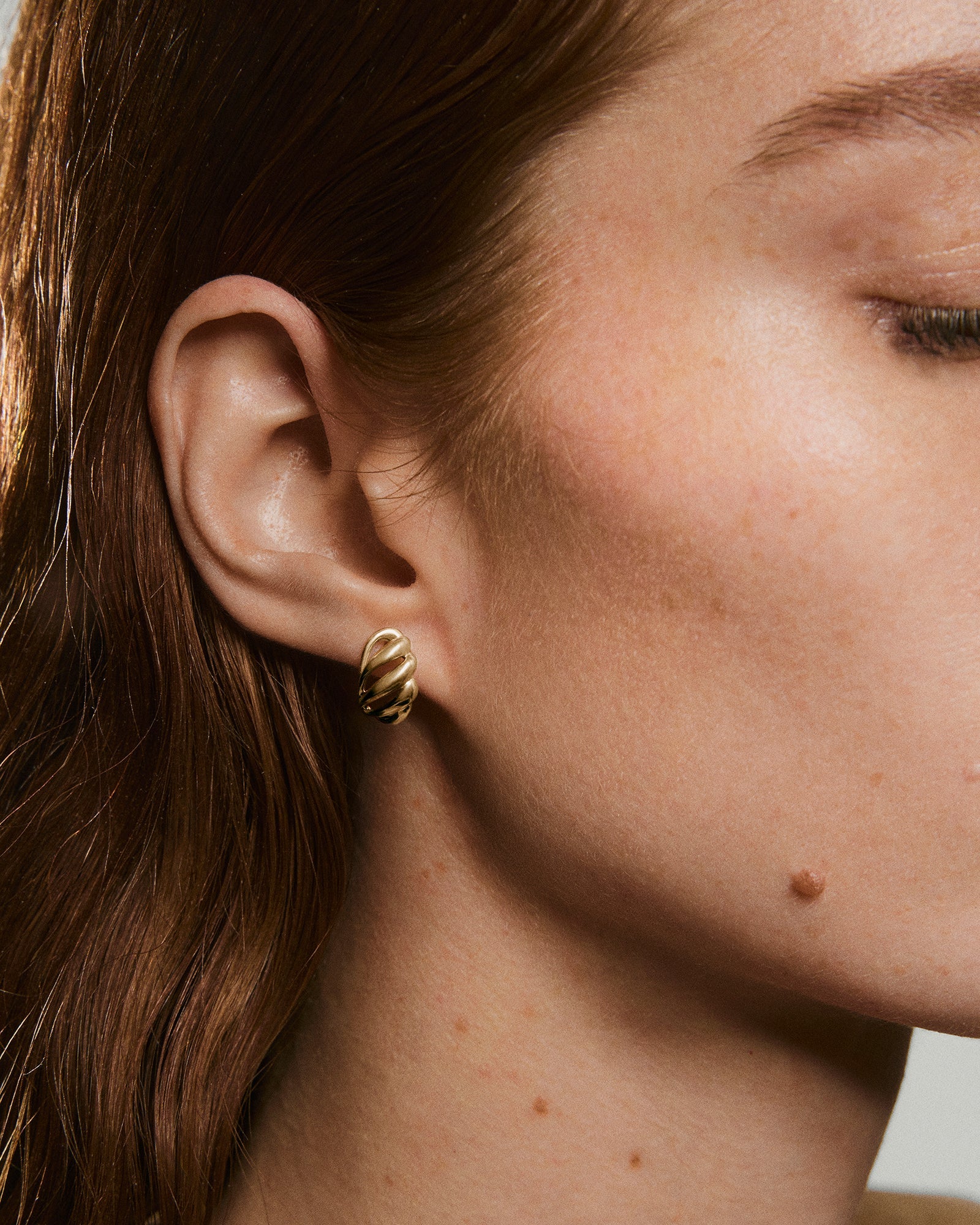 Close-up of a left ear with reddish-brown hair, featuring Bold Open DÃ´me Huggiesâa small, twist-style gold hoop earring with an asymmetrical, textured design.