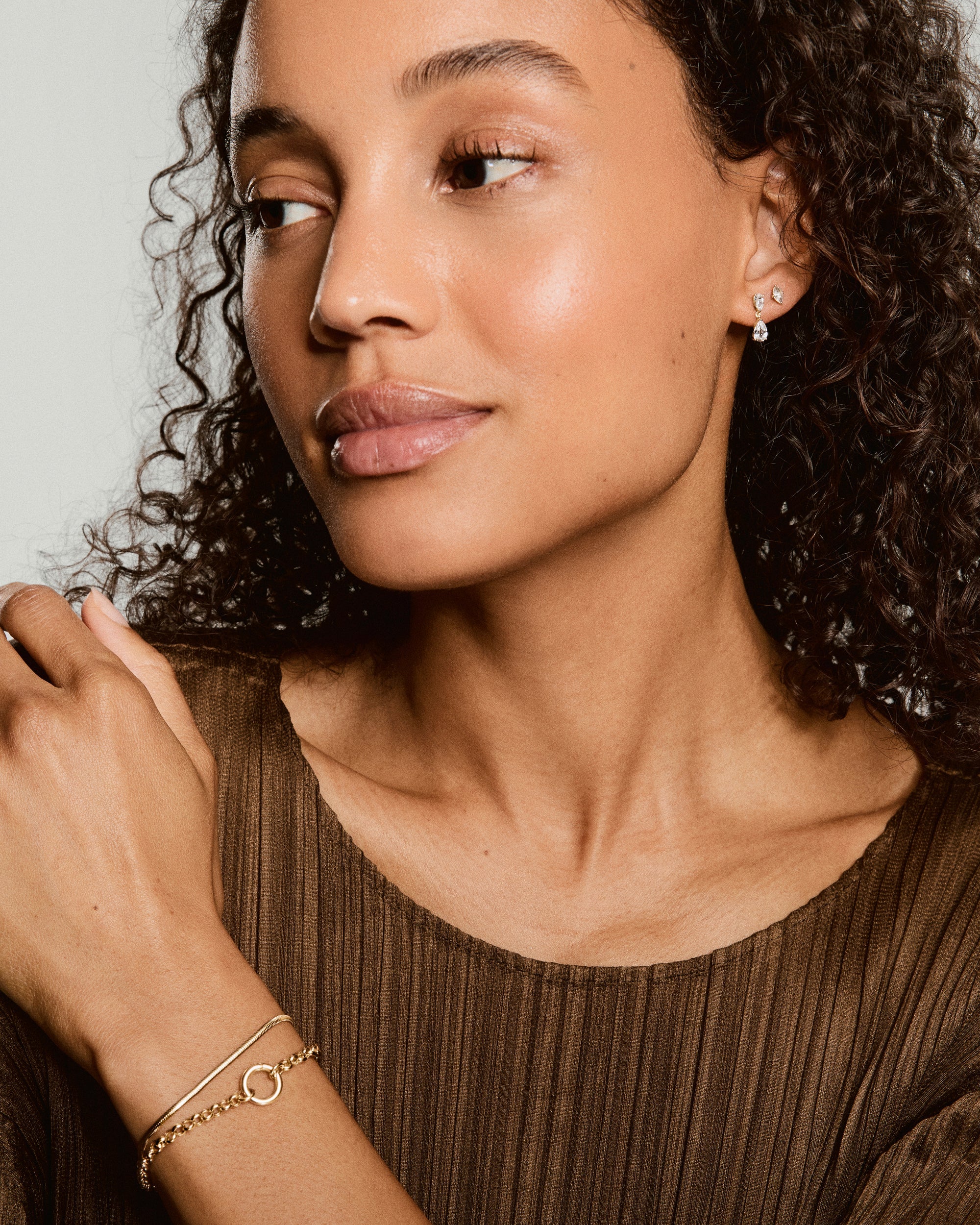 Young woman wearing Ada Double Pear Drop Earrings, featuring two pear-shaped gemstones. Earrings add shine and movement; complemented by gold bracelets on wrist.