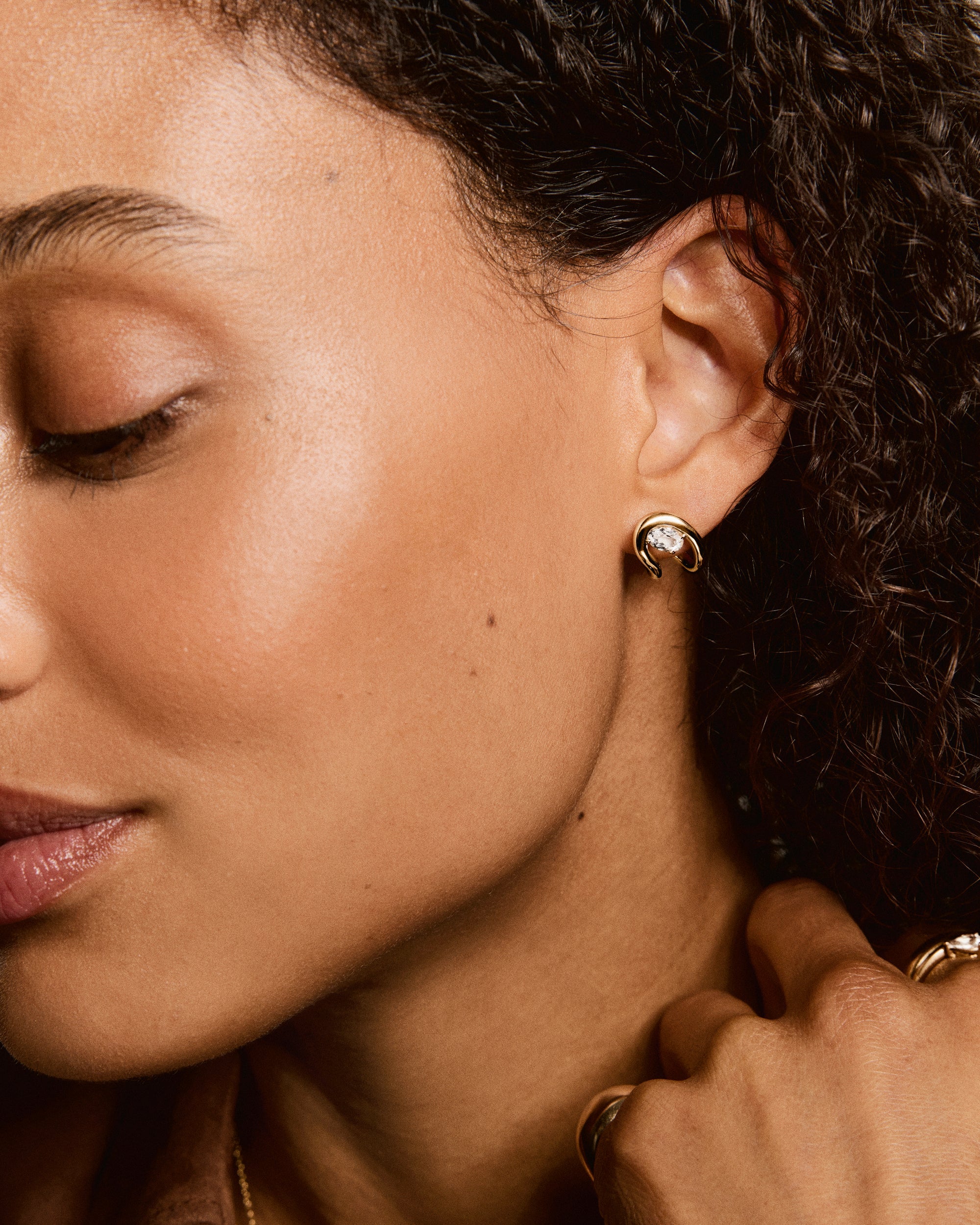 Close-up of woman wearing Organic DÃ´me Curve Gemstone Huggies; polished gold earring with clear gemstone, complemented by gold ring and fine chain necklace.