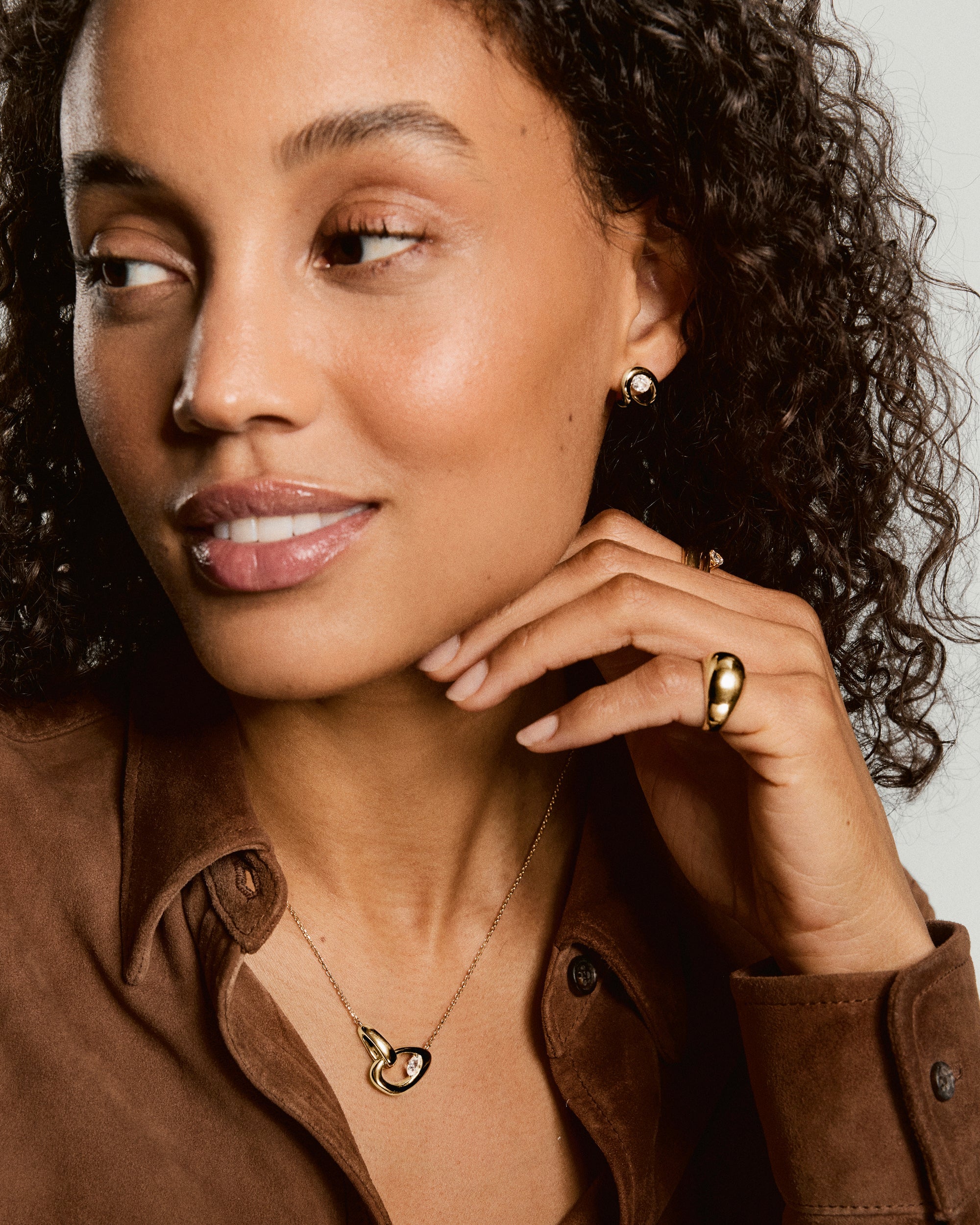 Woman wearing Organic DÃ´me Curve Gemstone Necklace with interlocking gold loops, one holding a faceted gem, complemented by gold hoop earrings and a chunky ring.