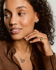 Woman wearing Organic DÃ´me Curve Gemstone Necklace with interlocking gold loops, one holding a faceted gem, complemented by gold hoop earrings and a chunky ring.
