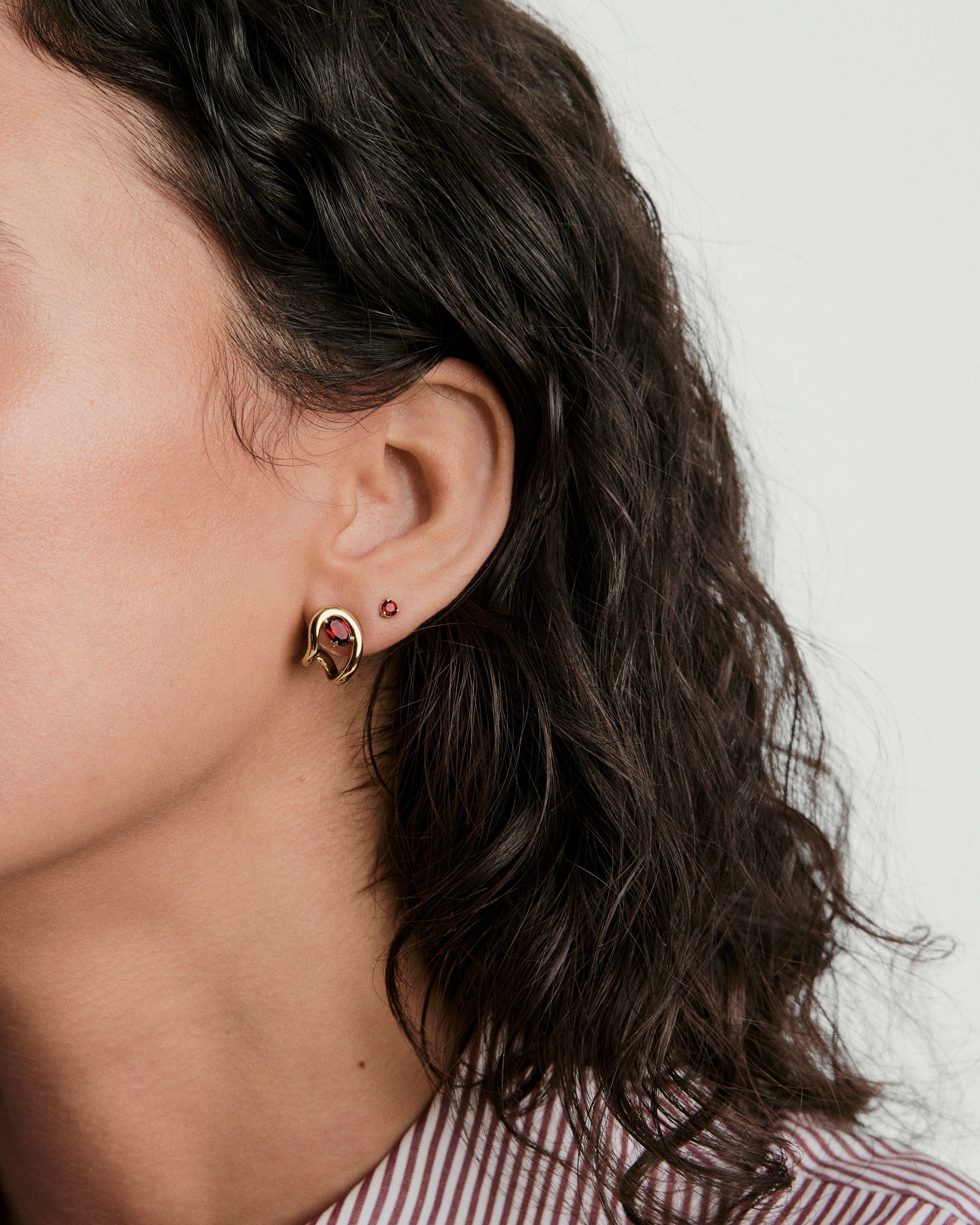 Organic DÃ´me Curve Gemstone Huggies showcased on a woman's ear, featuring a central red stone, highlighting the earring's artistic and unique design.