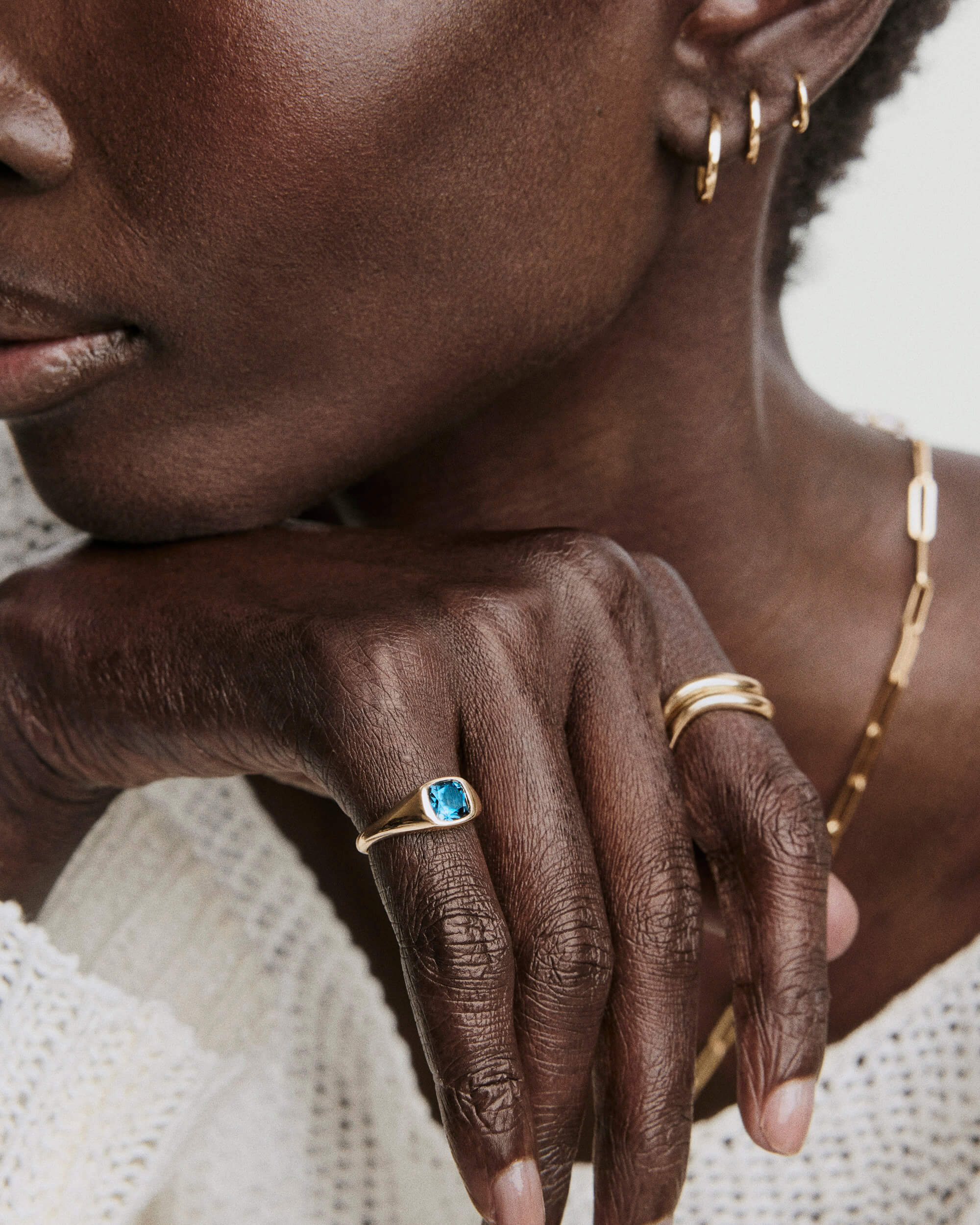 A close-up of a person wearing the Jude Pinky Signet Ring, featuring a blue gemstone on a gold band, paired with a gold necklace and earrings.