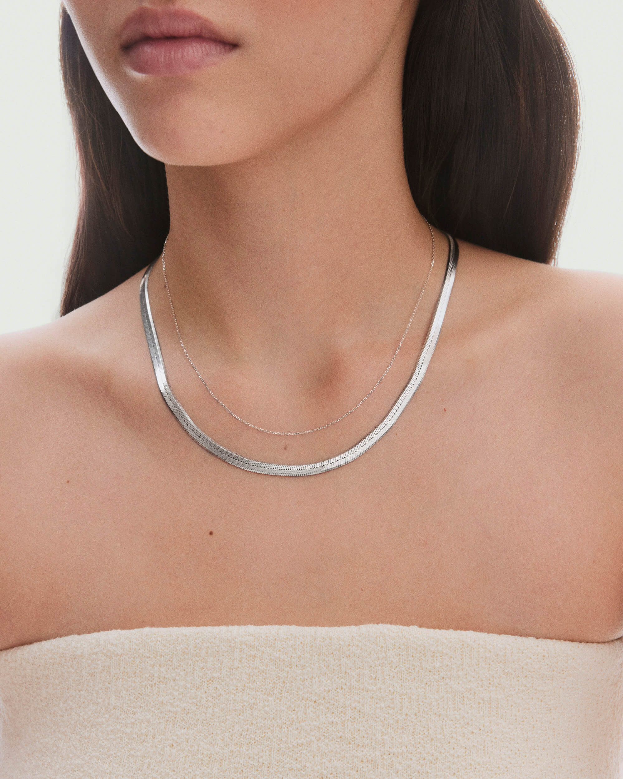 Bold Herringbone Chain Necklace layered with a delicate silver chain on a woman's neck, complementing a strapless beige top and dark hair.