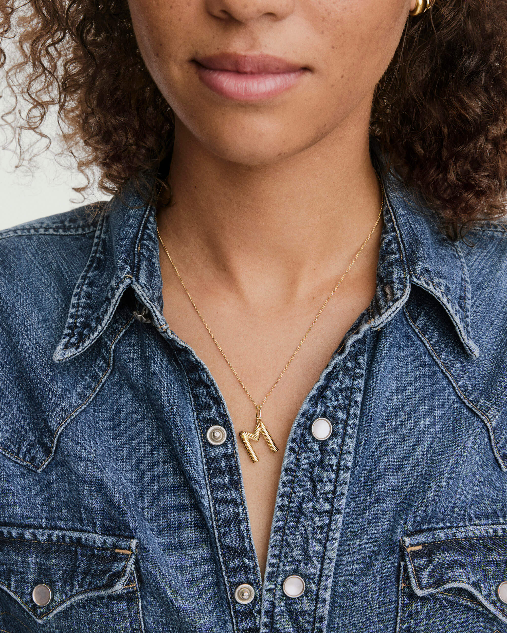 Close-up of a medium-wash denim shirt highlighting a Bold Letter Pendant Necklace, featuring an 18k gold vermeil M on a 20-22 inch chain.