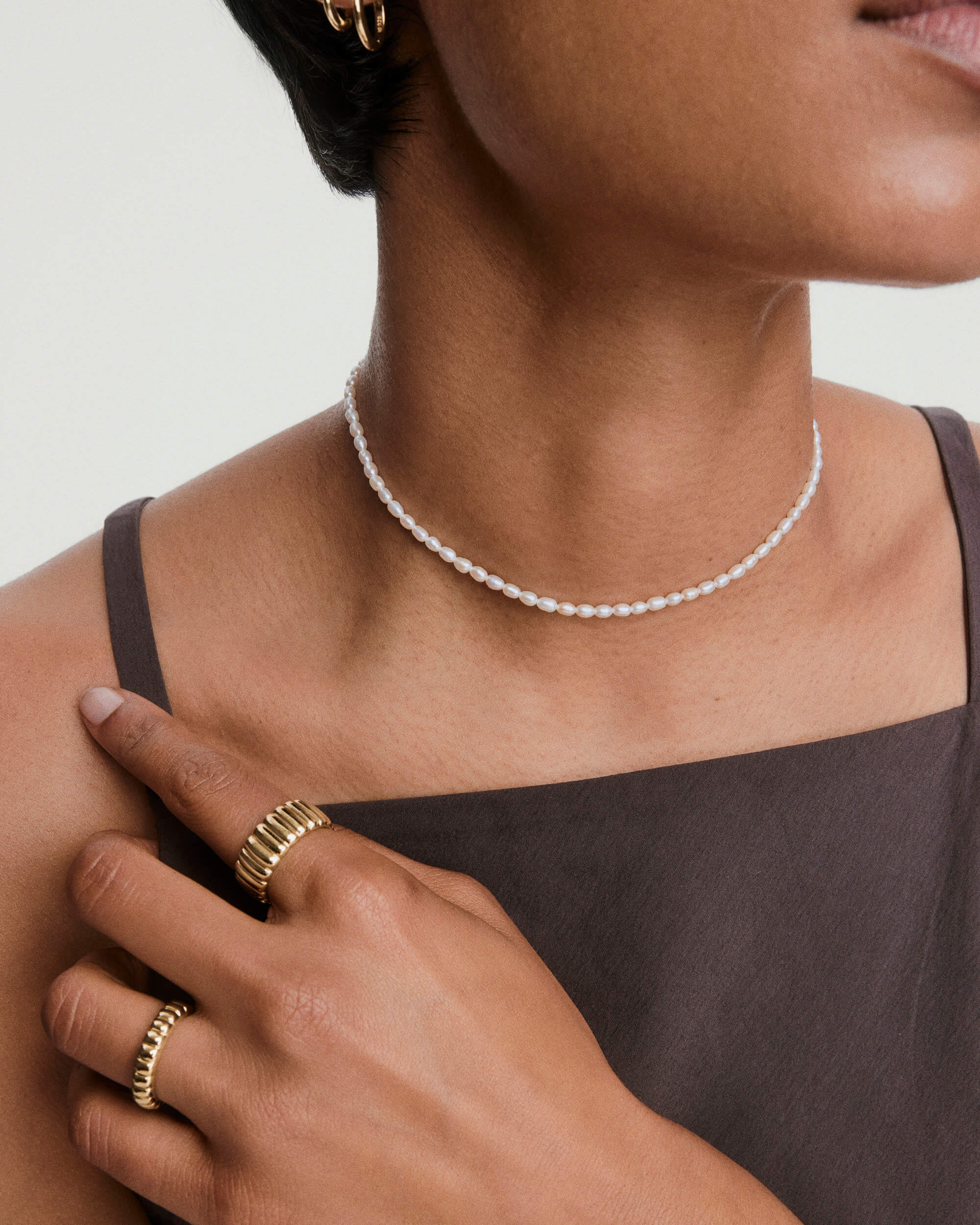 Tiny Pearl Necklace: A delicate choker of small white pearls is worn alongside gold rings, enhancing the elegance of a dark, sleeveless outfit.