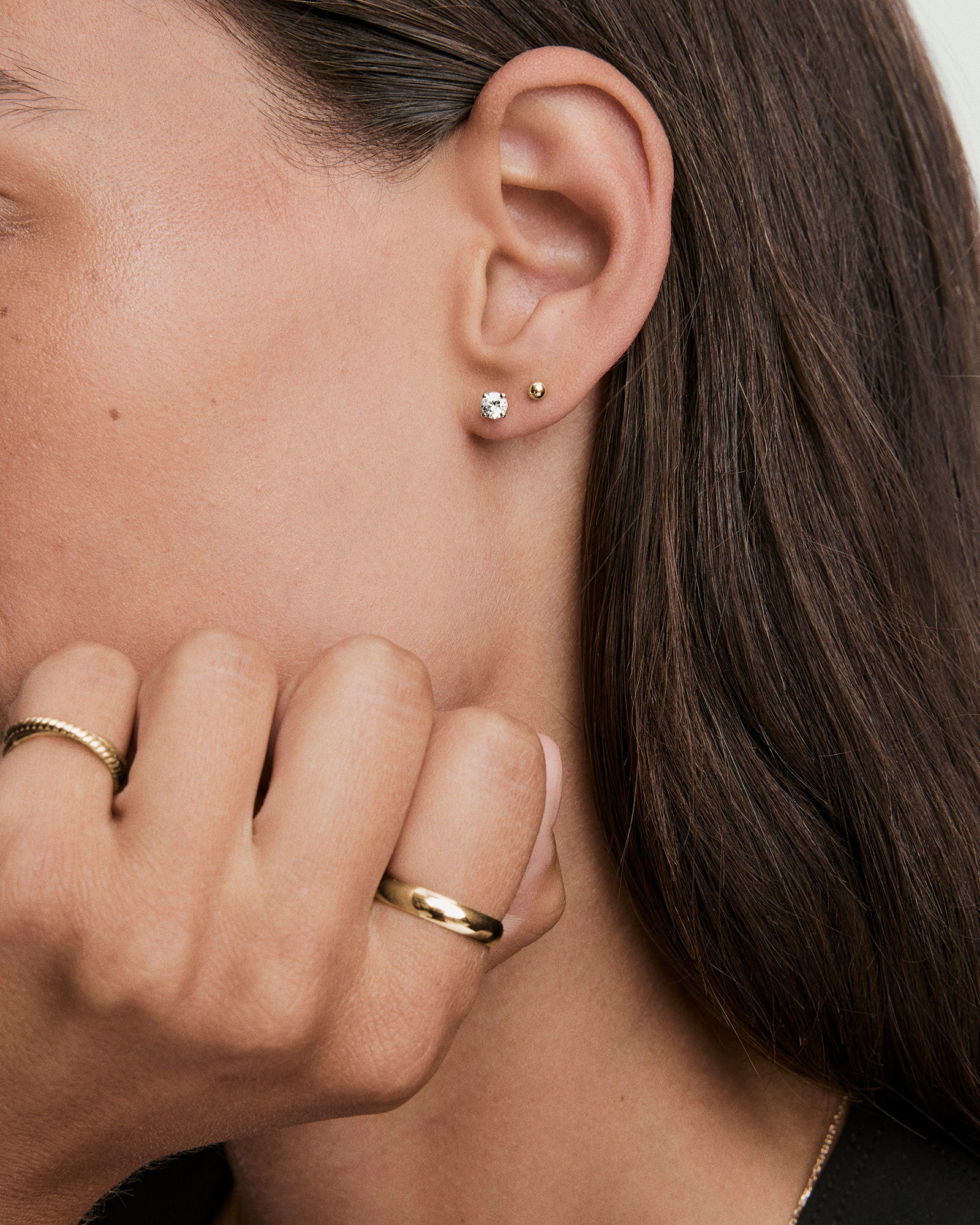 Side-profile of a woman's ear adorned with Round Lab Grown Sapphire Studs alongside a small gold ball, with gold rings on her hand.