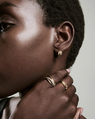 Close-up of a person's ear with Open DÃ´me Hoops, a small sculptural gold earring, paired with gold rings showcasing pavÃ© diamonds and a gemstone.