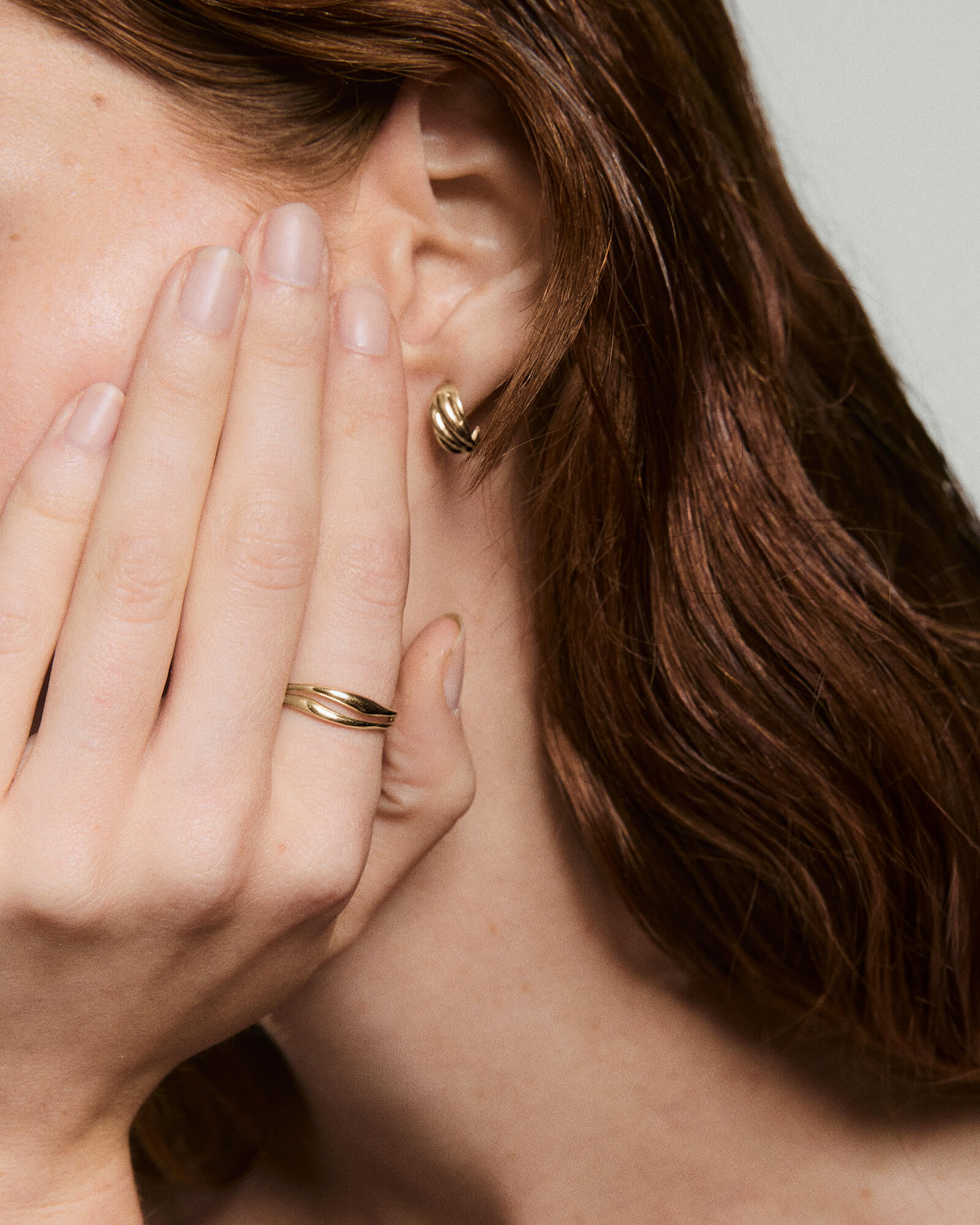 Close-up of a person wearing an Open DÃ´me Ring, a twisted gold band, on their ring finger, complementing a matching twisted gold hoop earring.