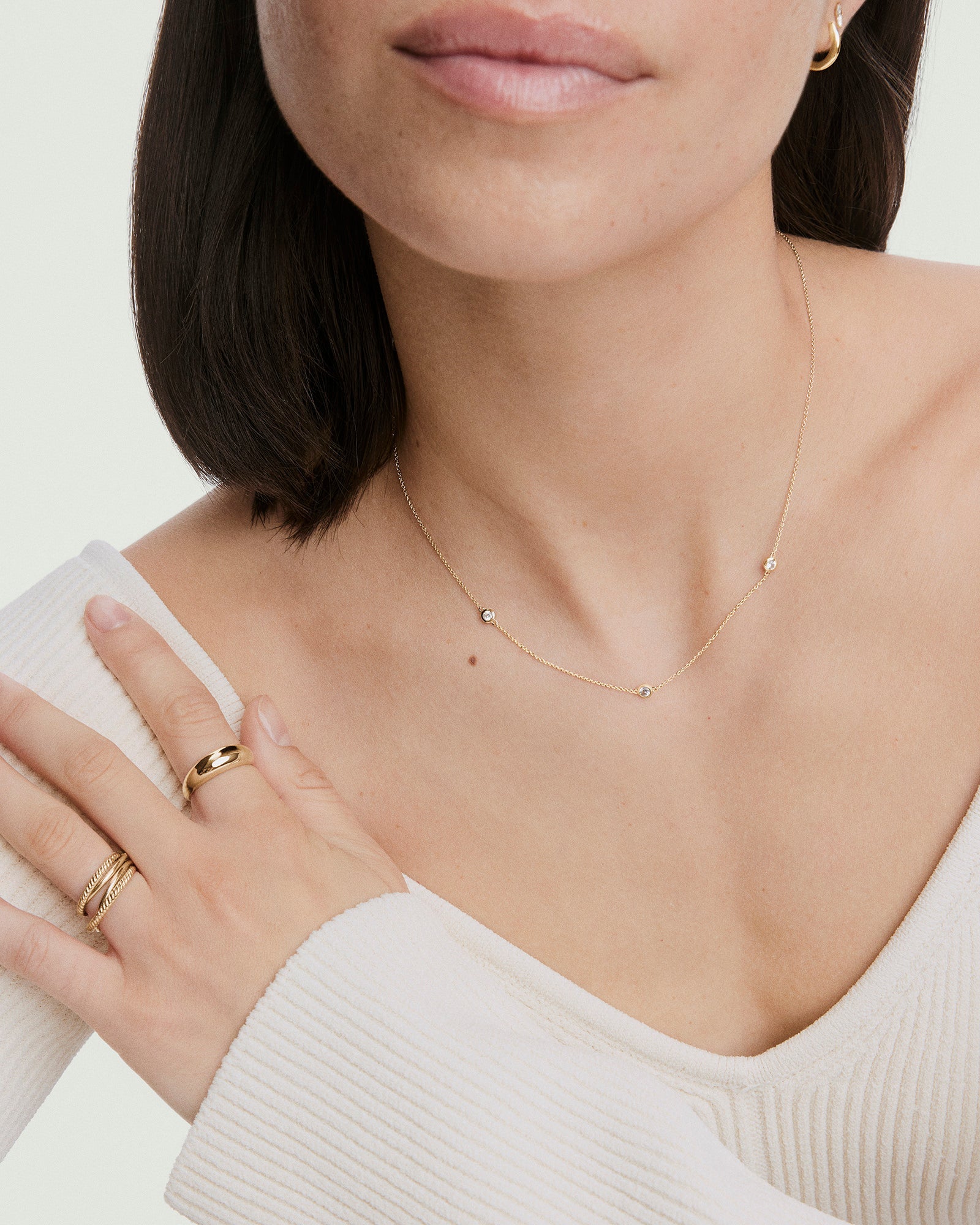 Woman wearing Satellite Necklace; a vermeil gold chain with three bezel-set white sapphires, paired with gold earrings and rings, complementing her off-white top.