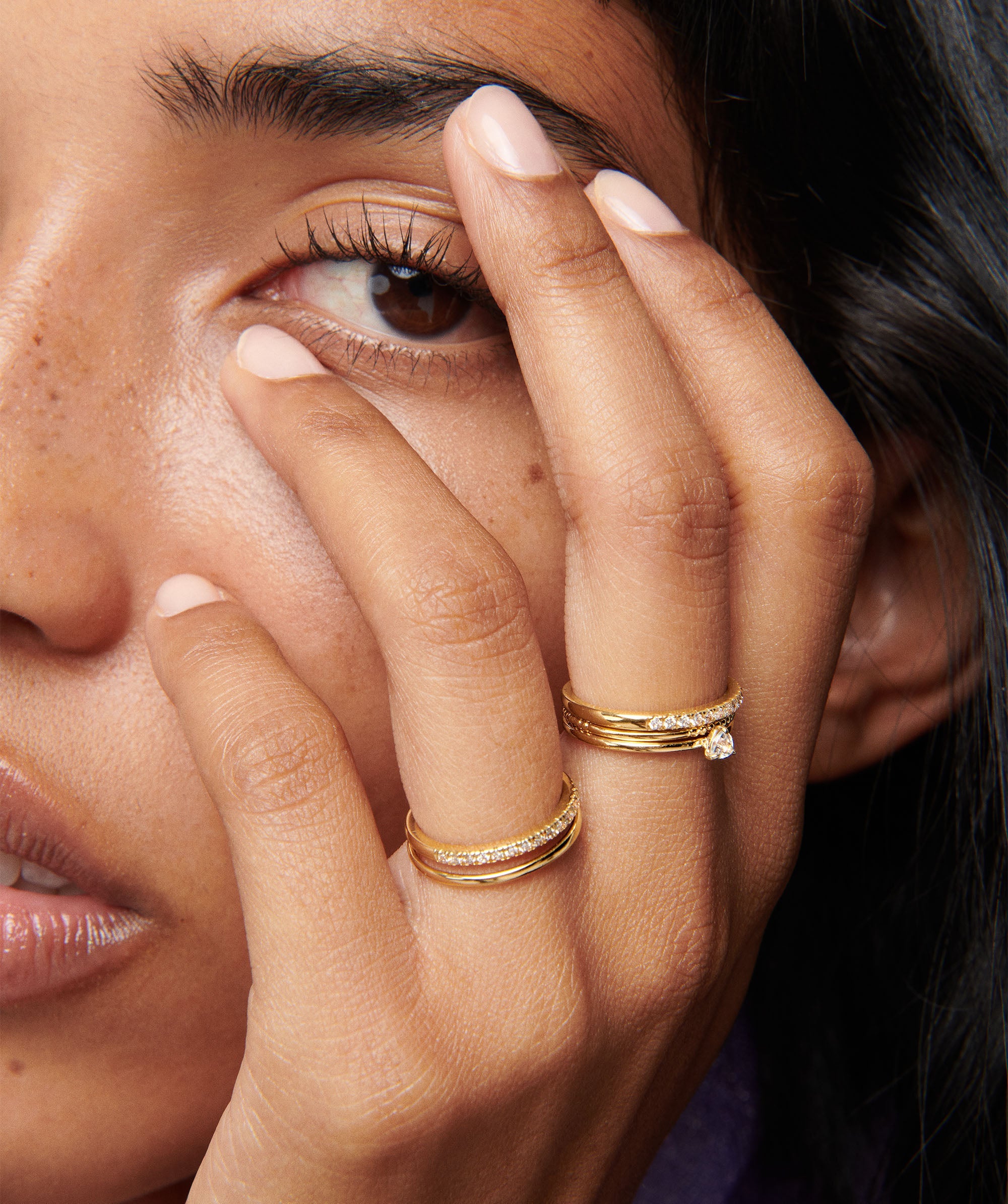 Close-up of a woman's face showcasing the Stacker Ring on her fingers. This essential piece is perfect for any collection.