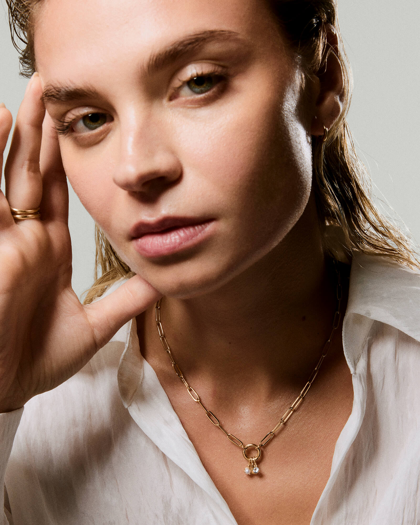Young woman wearing a Paperclip Chain Charm Necklace with a circular clasp and pearl-like charms, paired with slim gold rings and a small hoop earring.