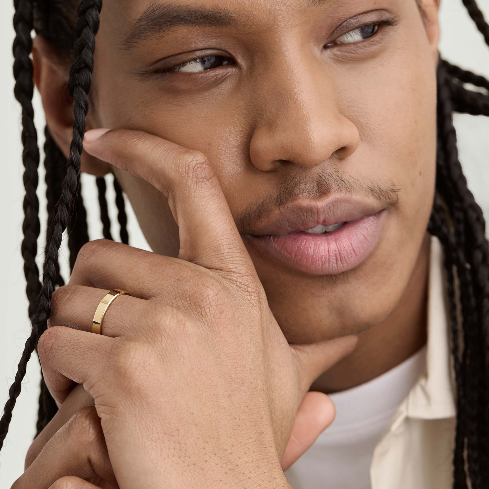 Man with braids holding his face, showcasing a 4mm Smooth Band gold bracelet on his wrist. The bracelet is lightweight and designed for lasting wear.