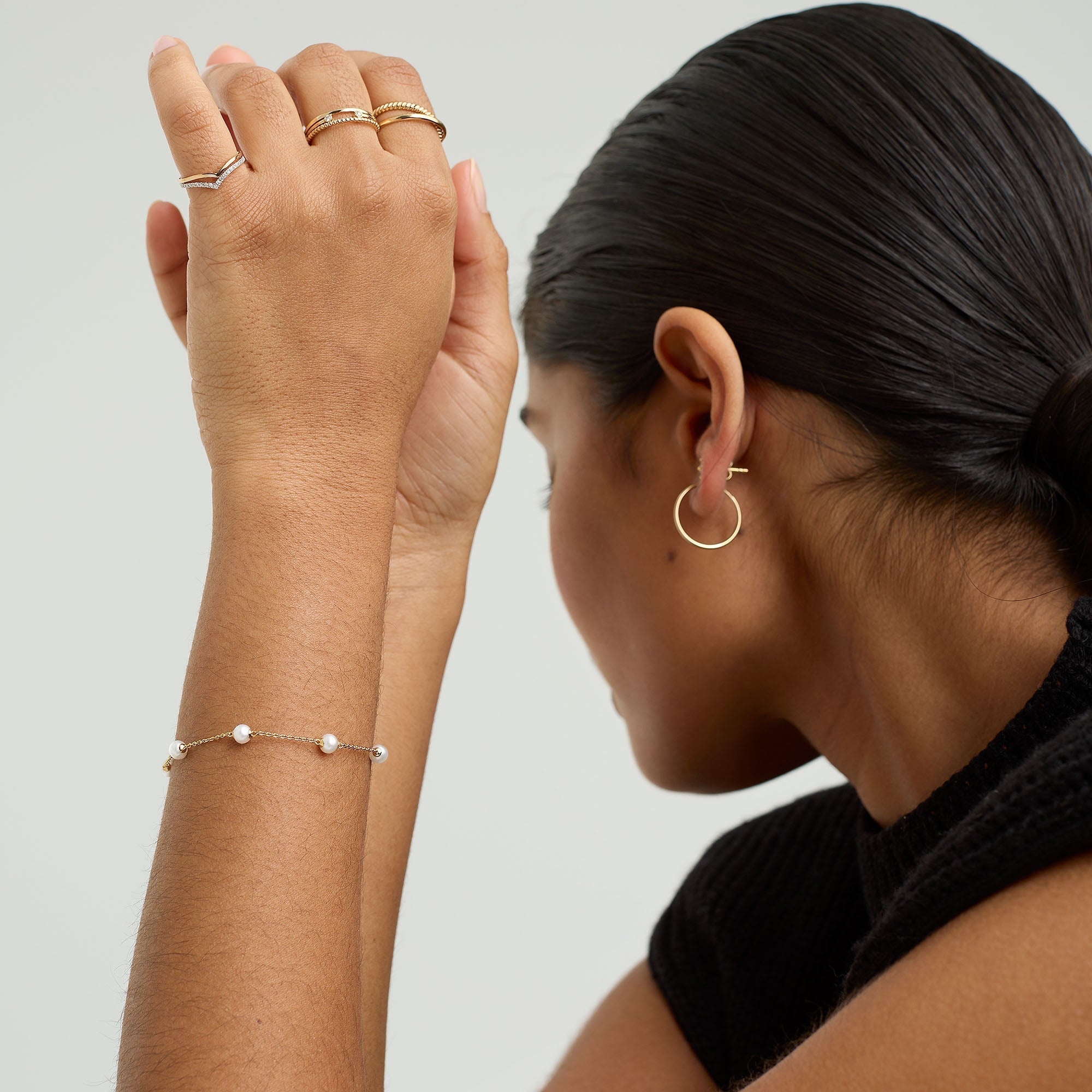 Woman's hand adorned with Mini Pearl Satellite Bracelet on a gold vermeil chain, complemented by gold rings and a close-up of her ear with a hoop earring.