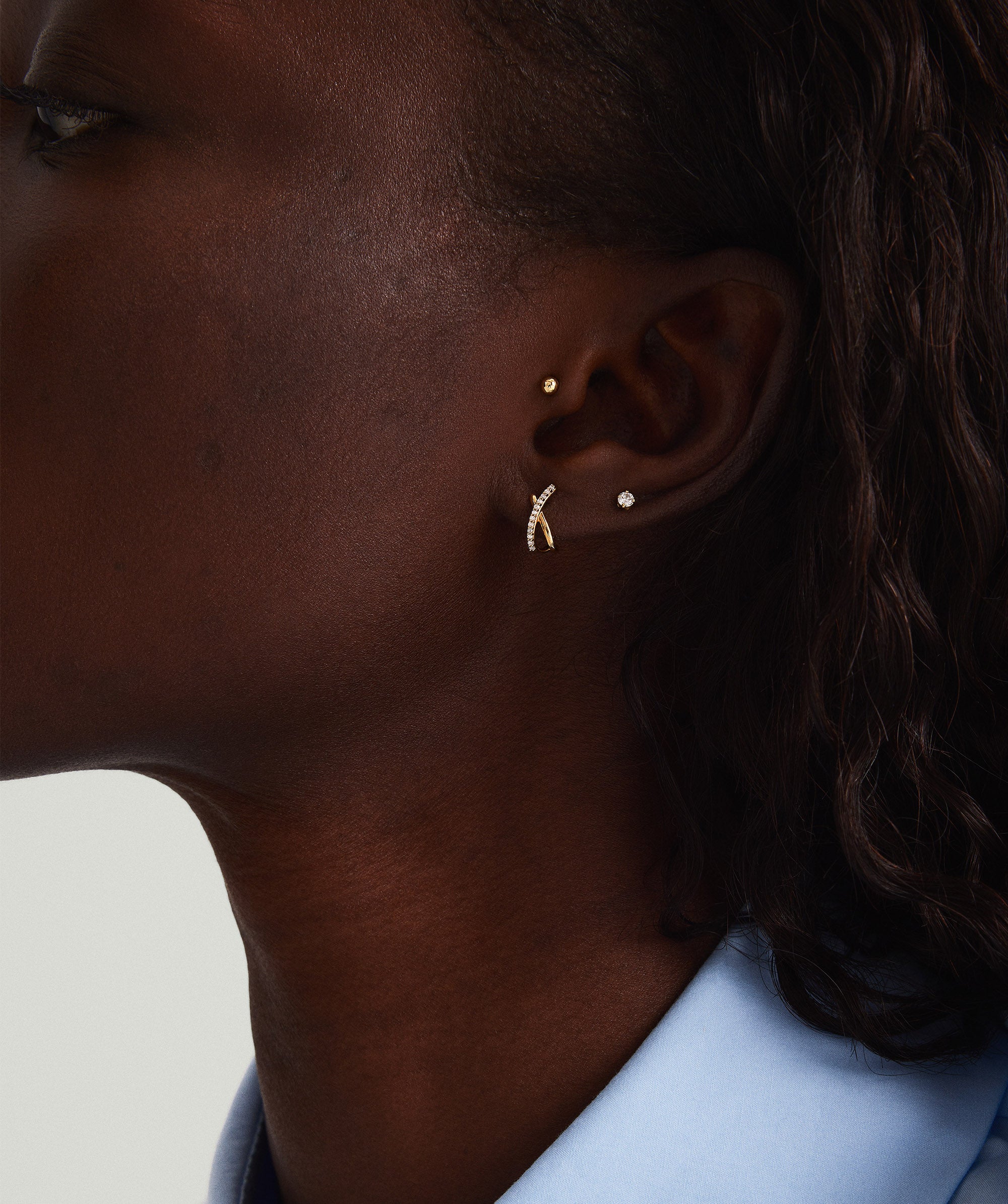 Close-up of a woman's ear wearing PavÃ© Diamond X Studs, handcrafted in 14k gold with pavÃ© diamonds, showcasing tubular lines overlapping gently.