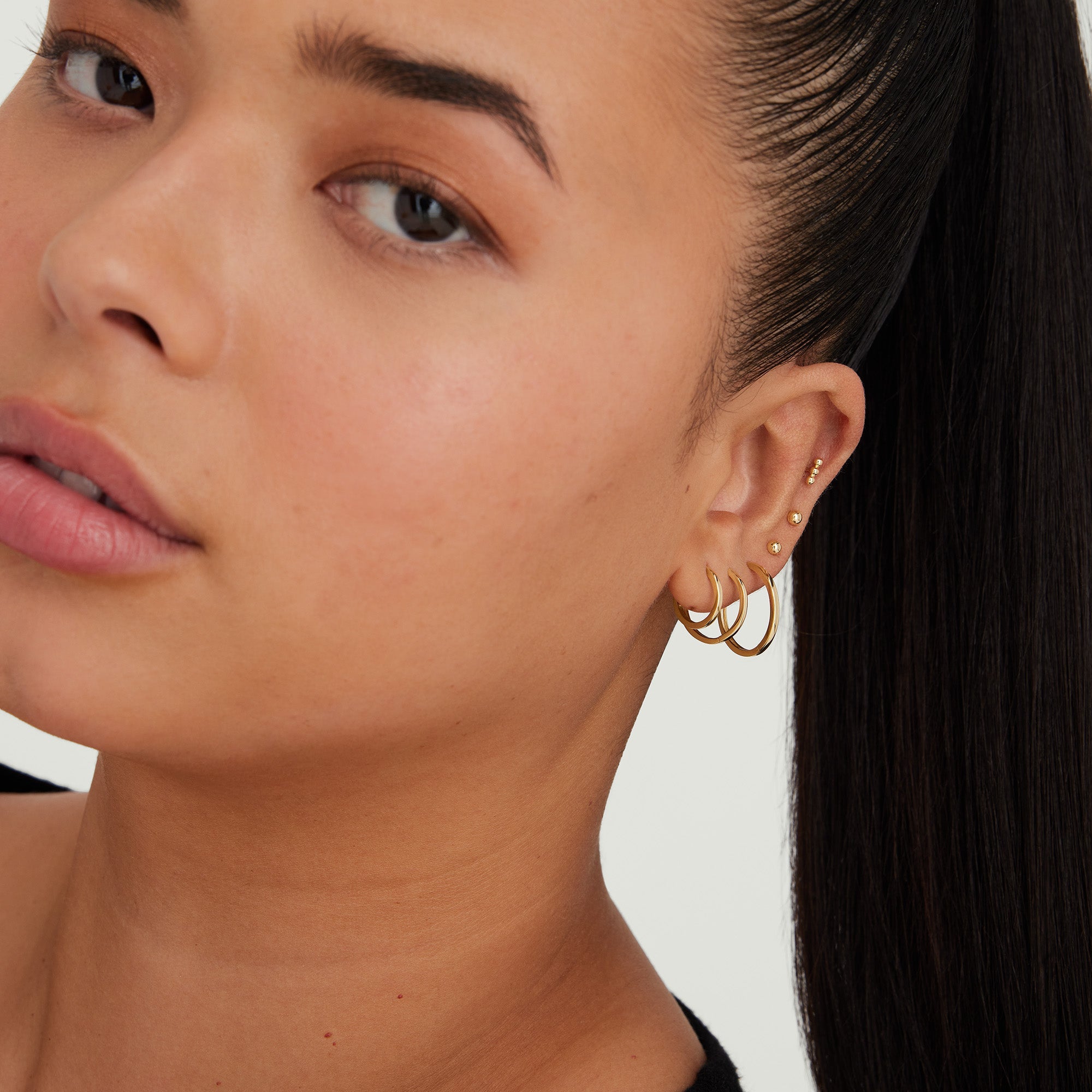 Close-up of a woman wearing Easy Small Hoops, showcasing multiple earrings on her ear. The handcrafted 14k gold hoops have a secure clicker closure.