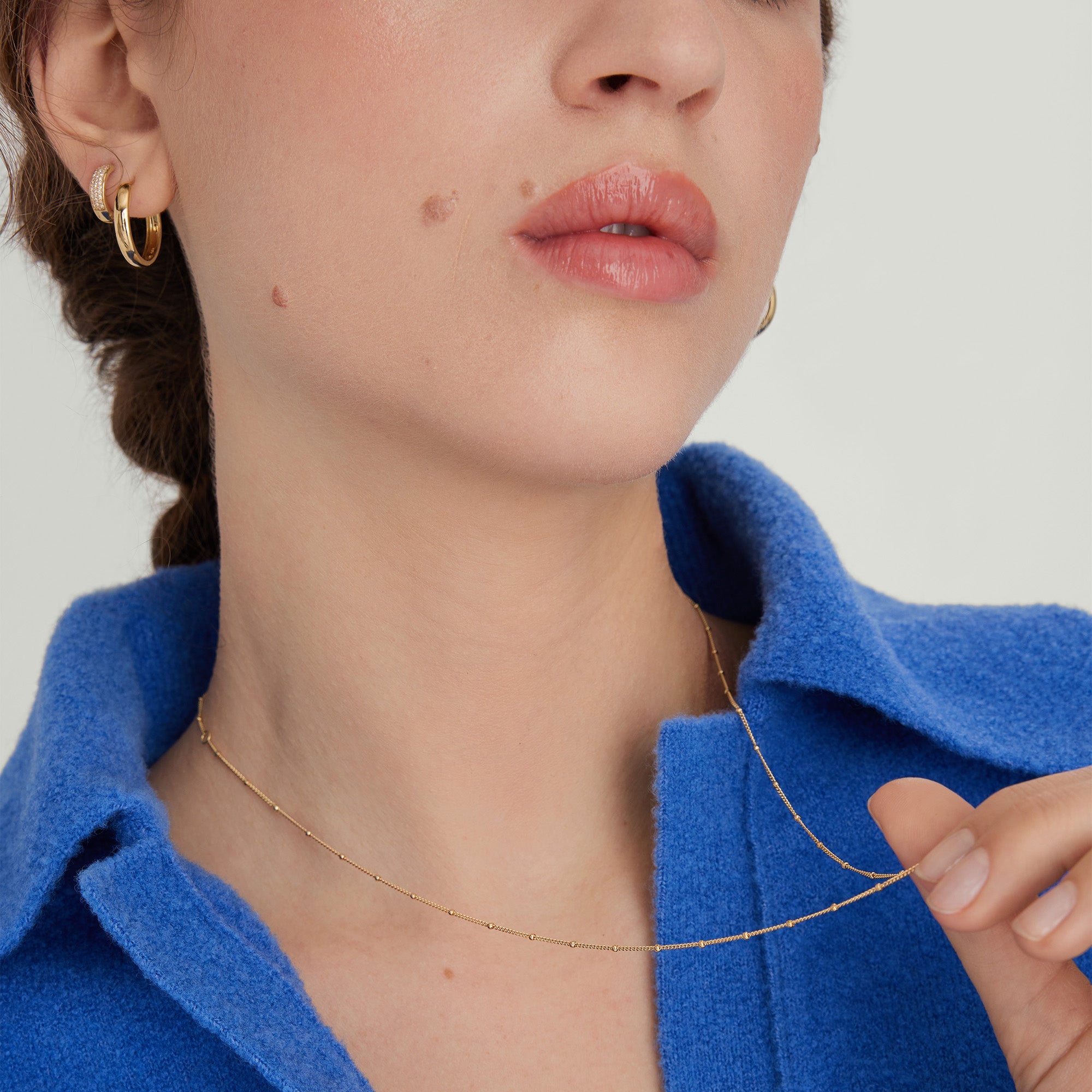Woman wearing a Spheres Chain Necklace with gold hoop earrings, blue shirt, and minimal makeup, showcasing the necklace's elegant and versatile style.
