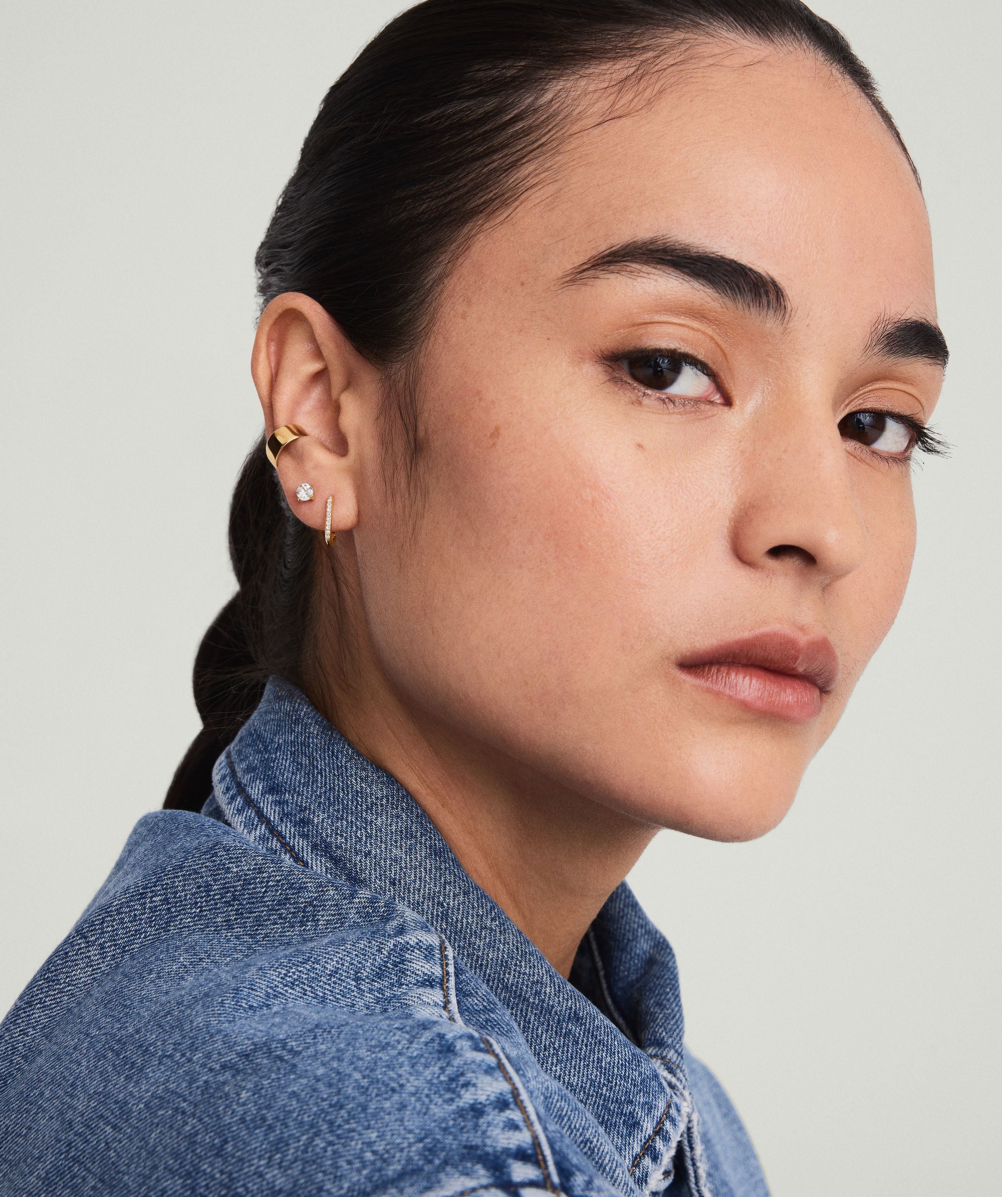 Woman wearing PavÃ© Diamond Huggie U Hoops, square-shaped, handcrafted in 14k solid gold with diamond accents, complementing her blue jacket.