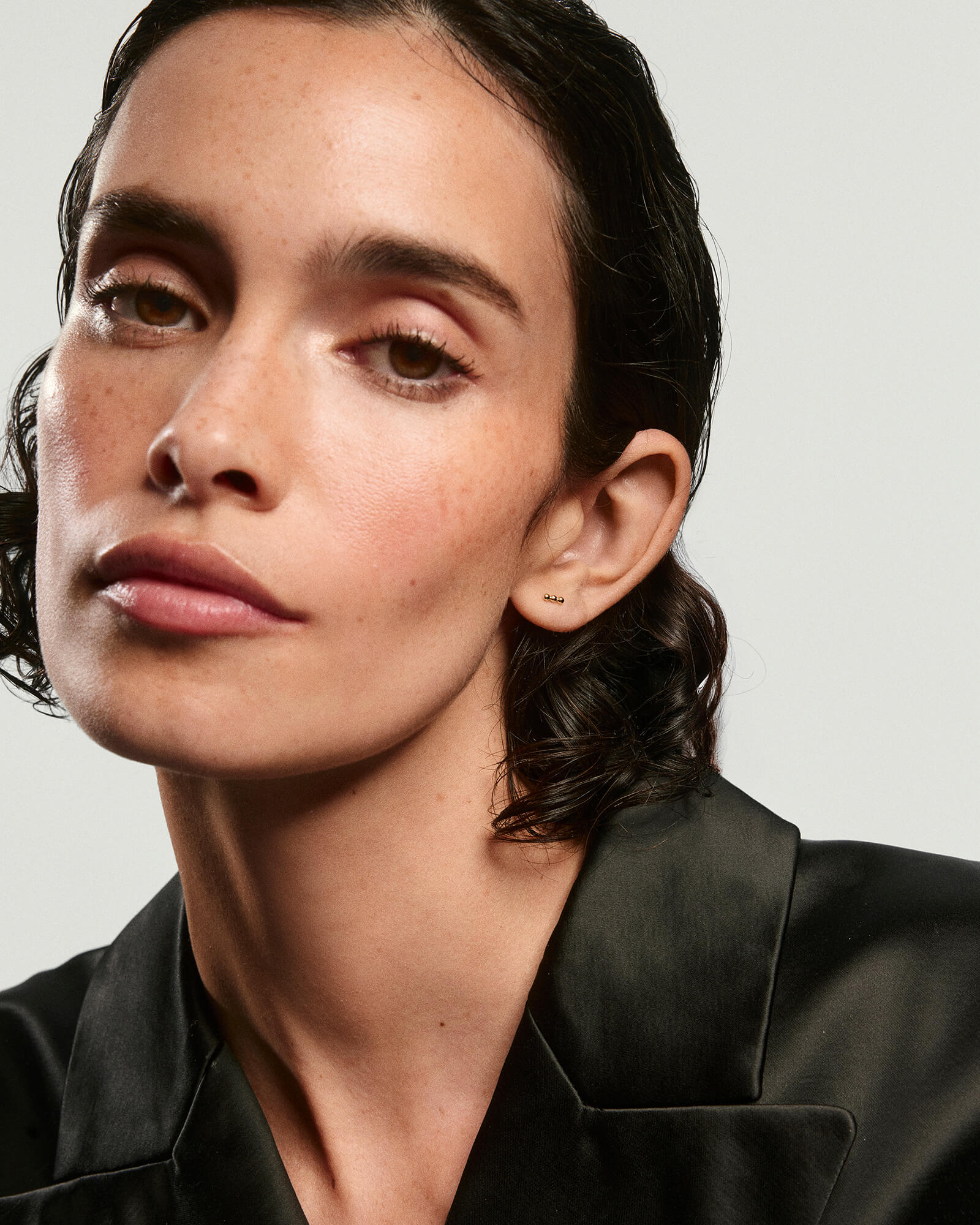 Young woman wearing Spheres Bar Studs, featuring a trio of tiny gold spheres, complemented by a clean, dewy complexion and dark blazer.