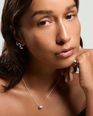 Young woman wearing the Lucia Necklace, featuring an oval bezel-set lab-grown sapphire on a chain, complemented by silver hoop earrings and a chunky ring.