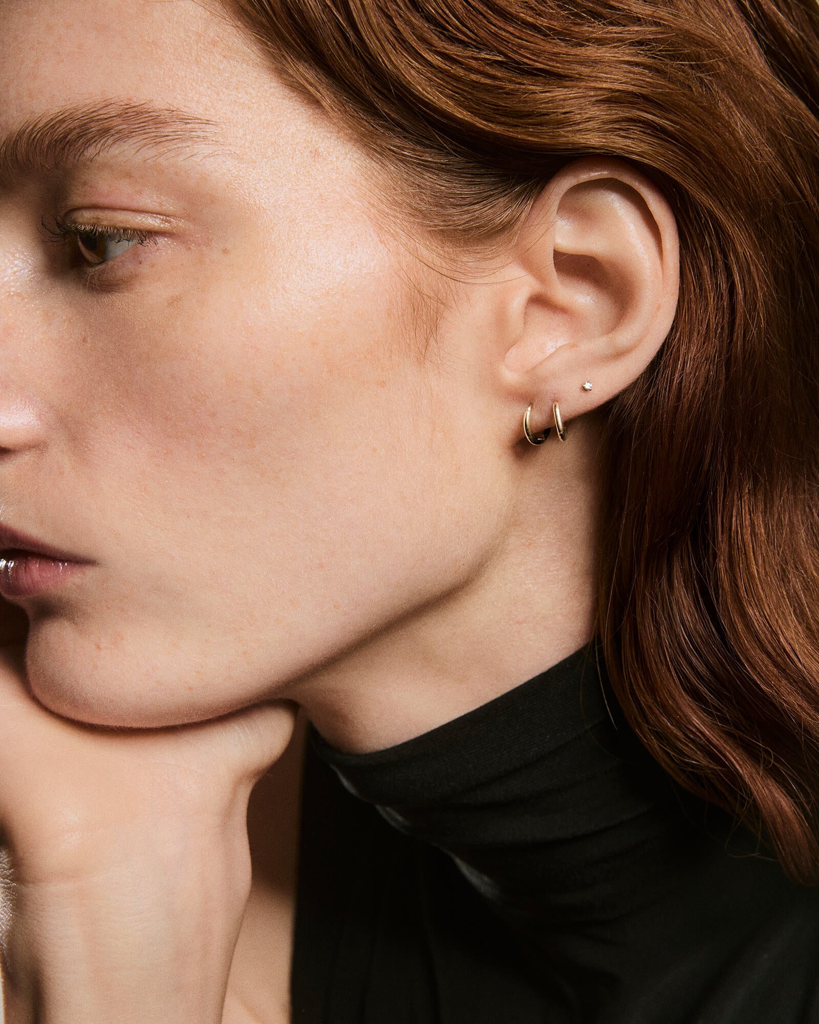 Close-up of a person's ear wearing Piercing Studio - Baby Diamond Stud, a small, elegant stud earring with a brilliant diamond, above two gold hoops.