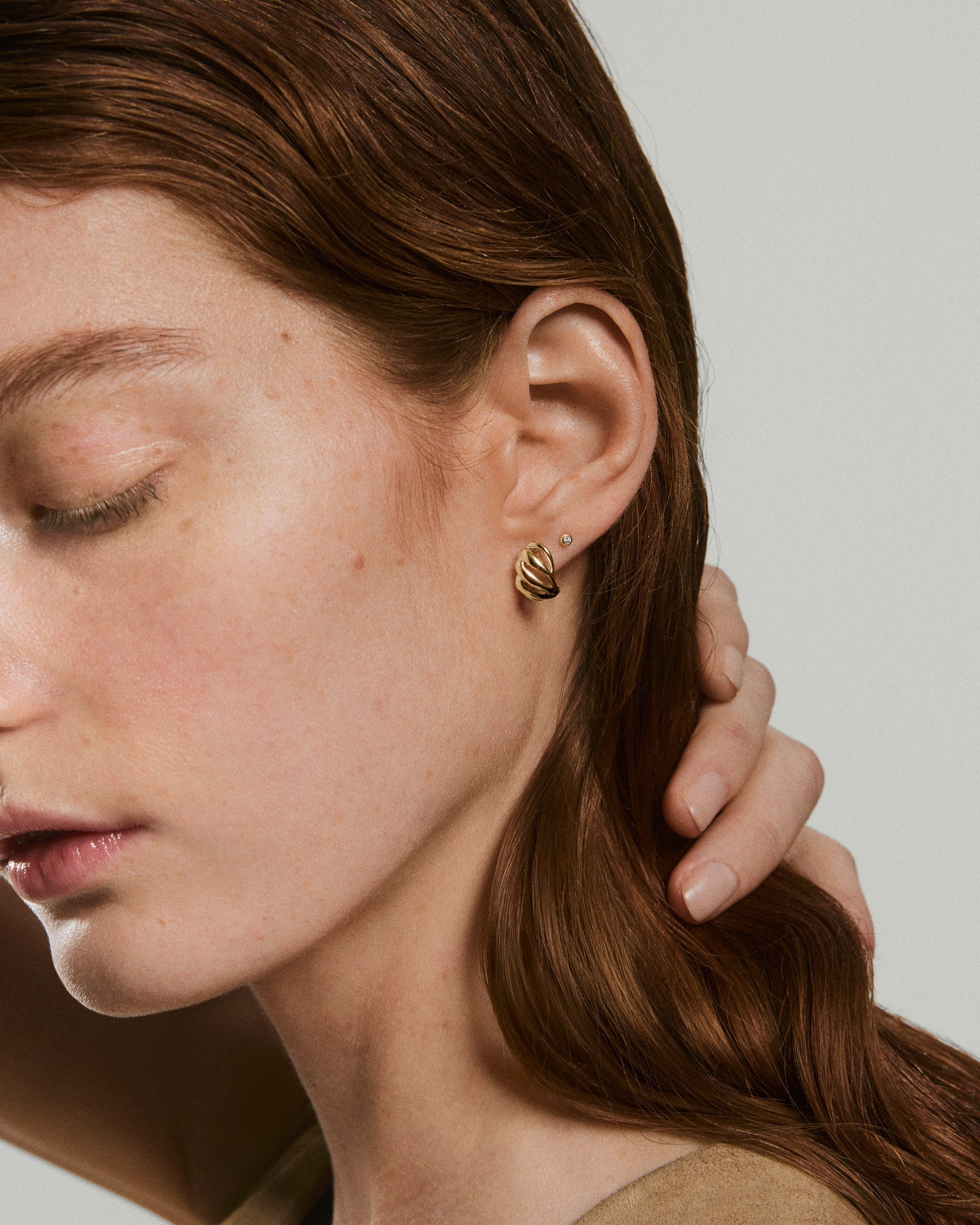 Young woman wearing Bold Open DÃ´me Huggies, featuring a sculptural, twisted gold hoop design, alongside a small stud earring, highlighting their textured, asymmetrical style.