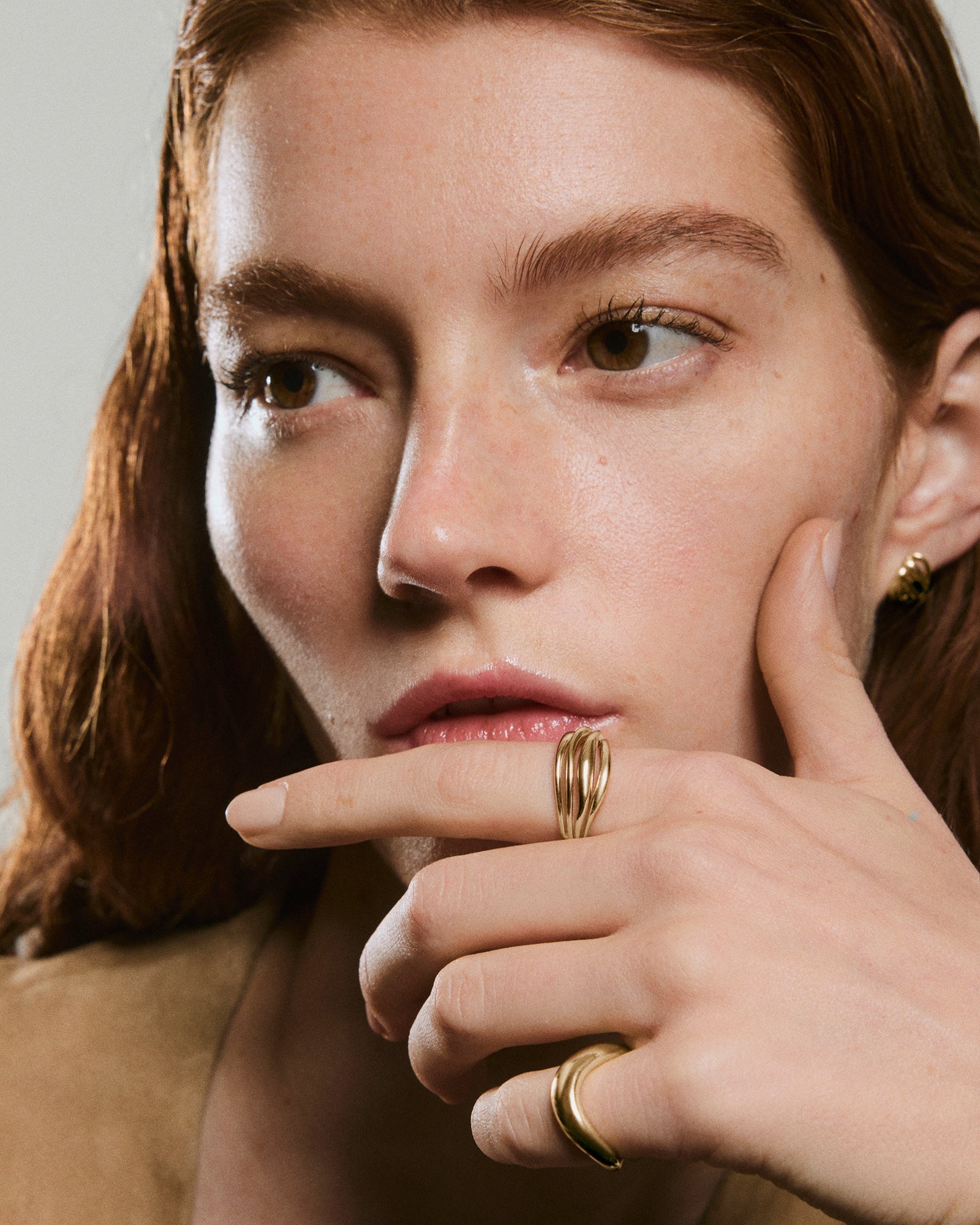 Young woman with auburn hair showcases a Bold Open DÃ´me Ring on her index finger, a gold sculptural band highlighting its open space and asymmetrical design.