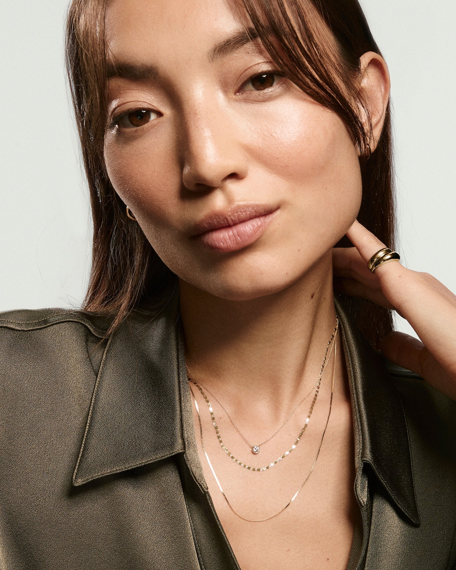 Young woman wearing the Mini Serpentine Necklace, featuring delicate S-curve links, layered with other gold necklaces, and a chunky gold ring on her hand.